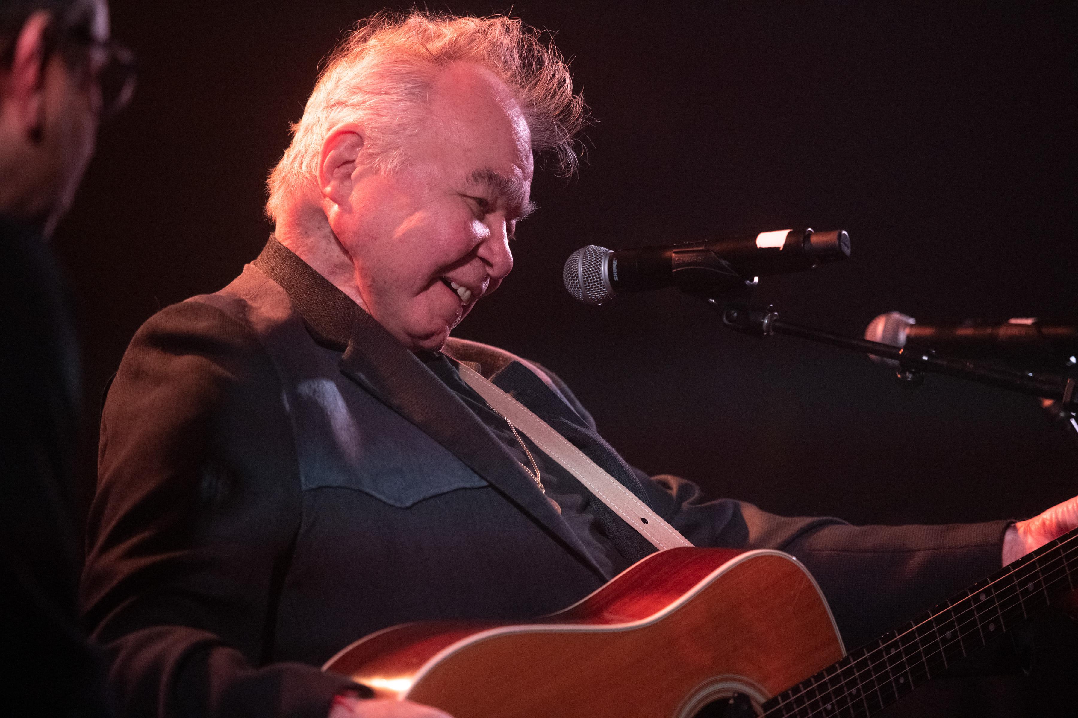 John Prine, smiling, plays an acoustic guitar onstage at the Troubadour.