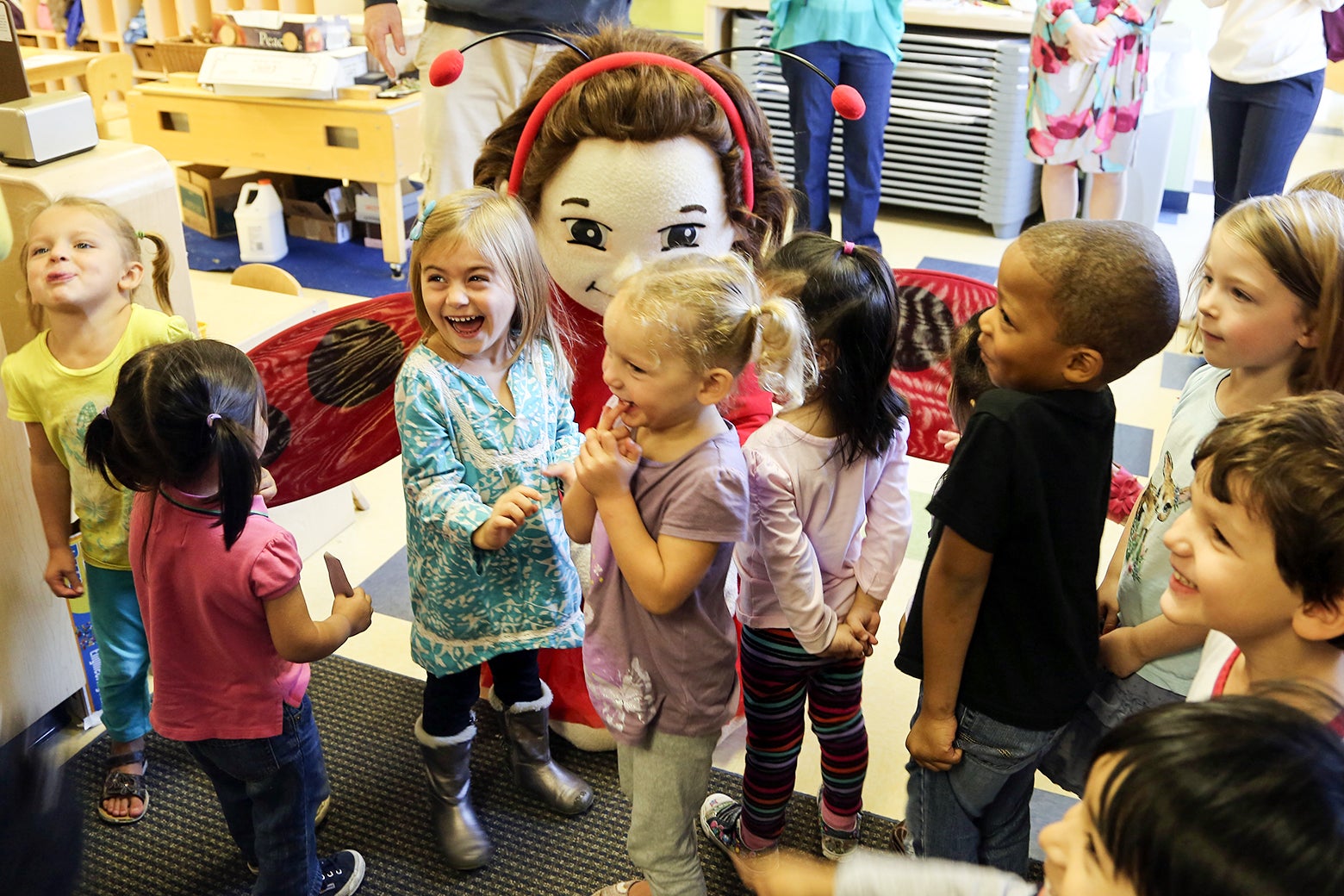Laughing preschoolers gather around a character dressed as Ladybug Girl in a schoolroom setting.