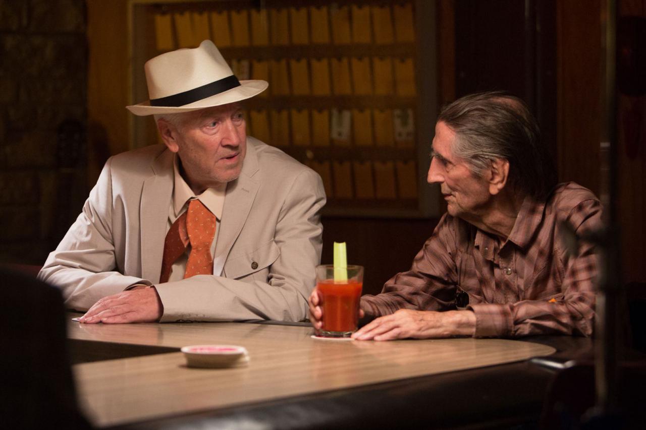 Two men sit at a bar, one in a beige suit and a hat and the other in a button-down shirt.