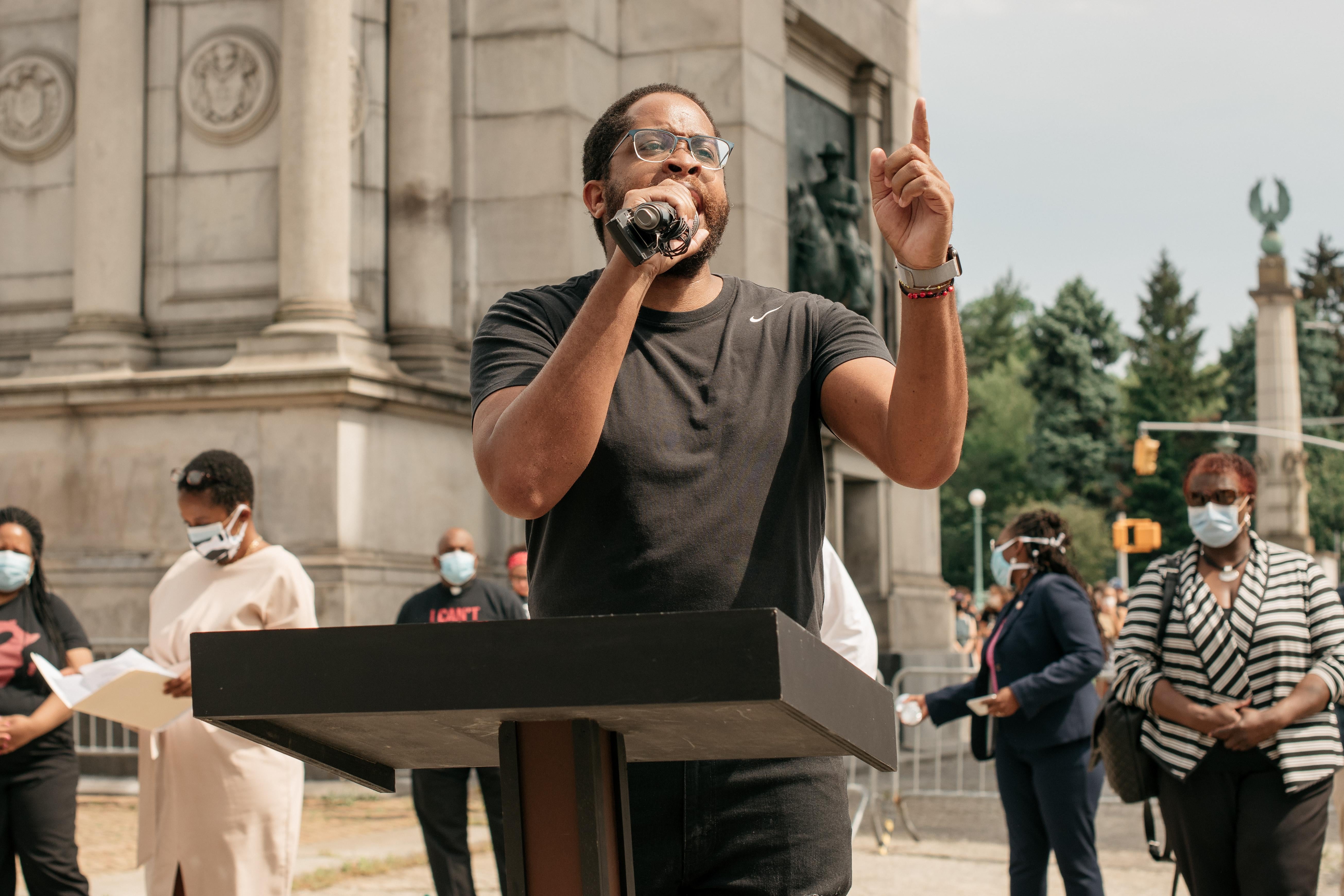 Zellnor Myrie stands at a lectern and speaks into a microphone, with people in face masks behind him