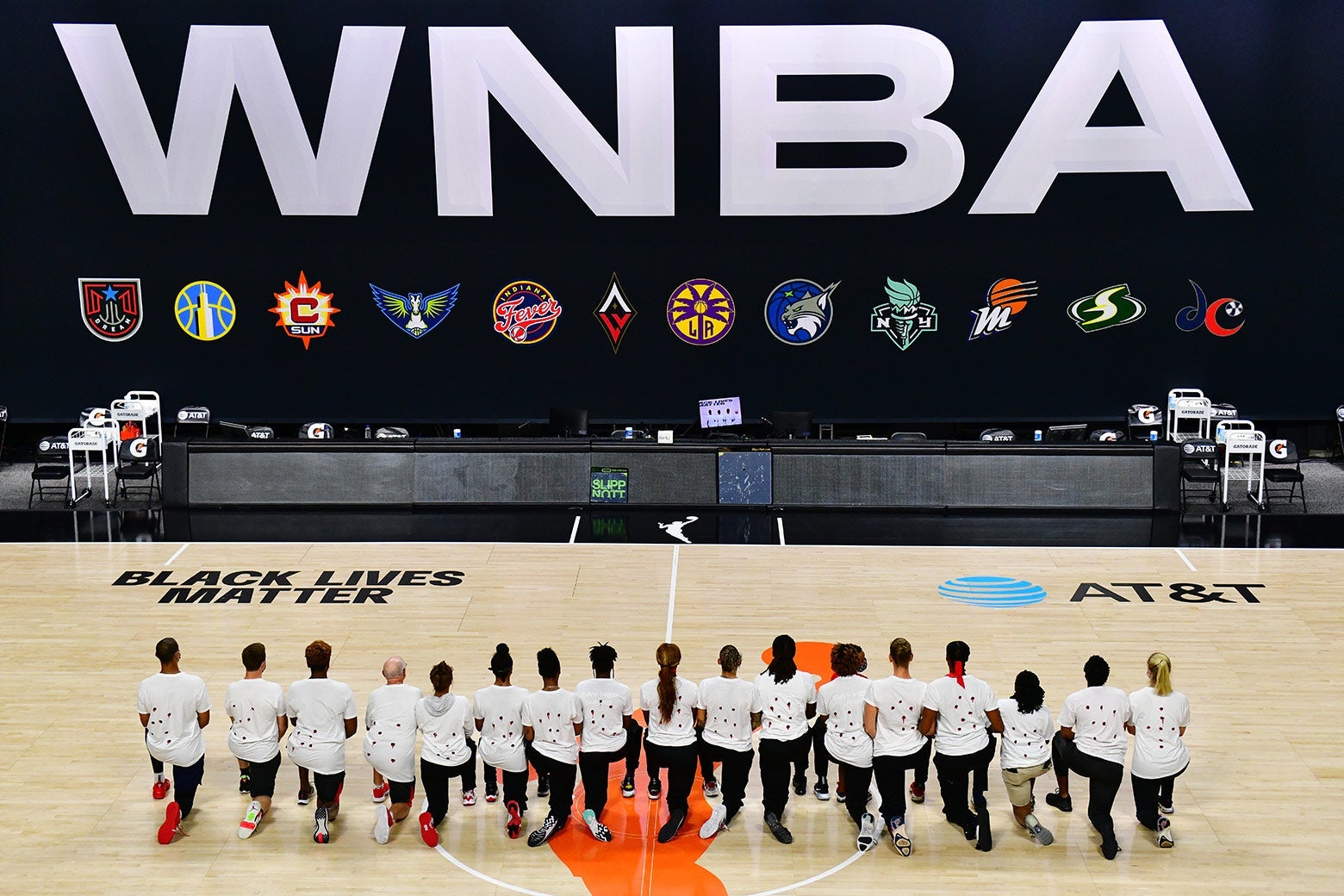 Mystics players wearing protest shirts kneel in a line on a basketball court in front of a big WNBA sign