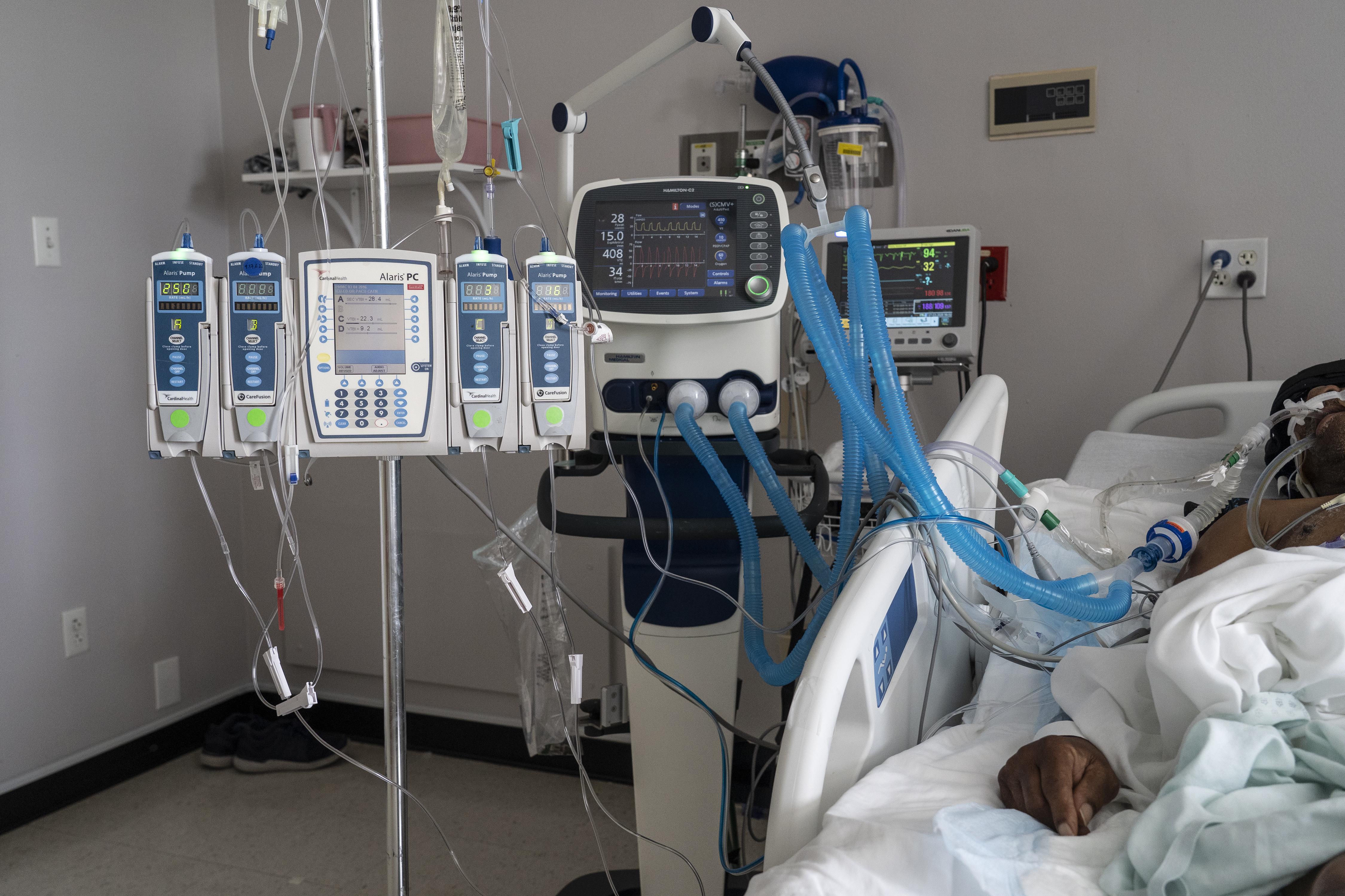 Hospital machinery and a ventilator stand to the left of a hospital bed. On the right, part of a patient's body is visible, connected to the machinery.