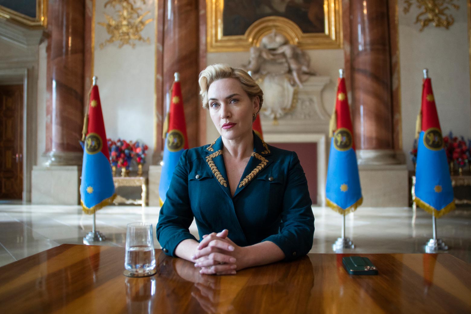 Kate Winslet as Chancellor Elena, wearing a blue power suit and sitting at a table with flags in the background.