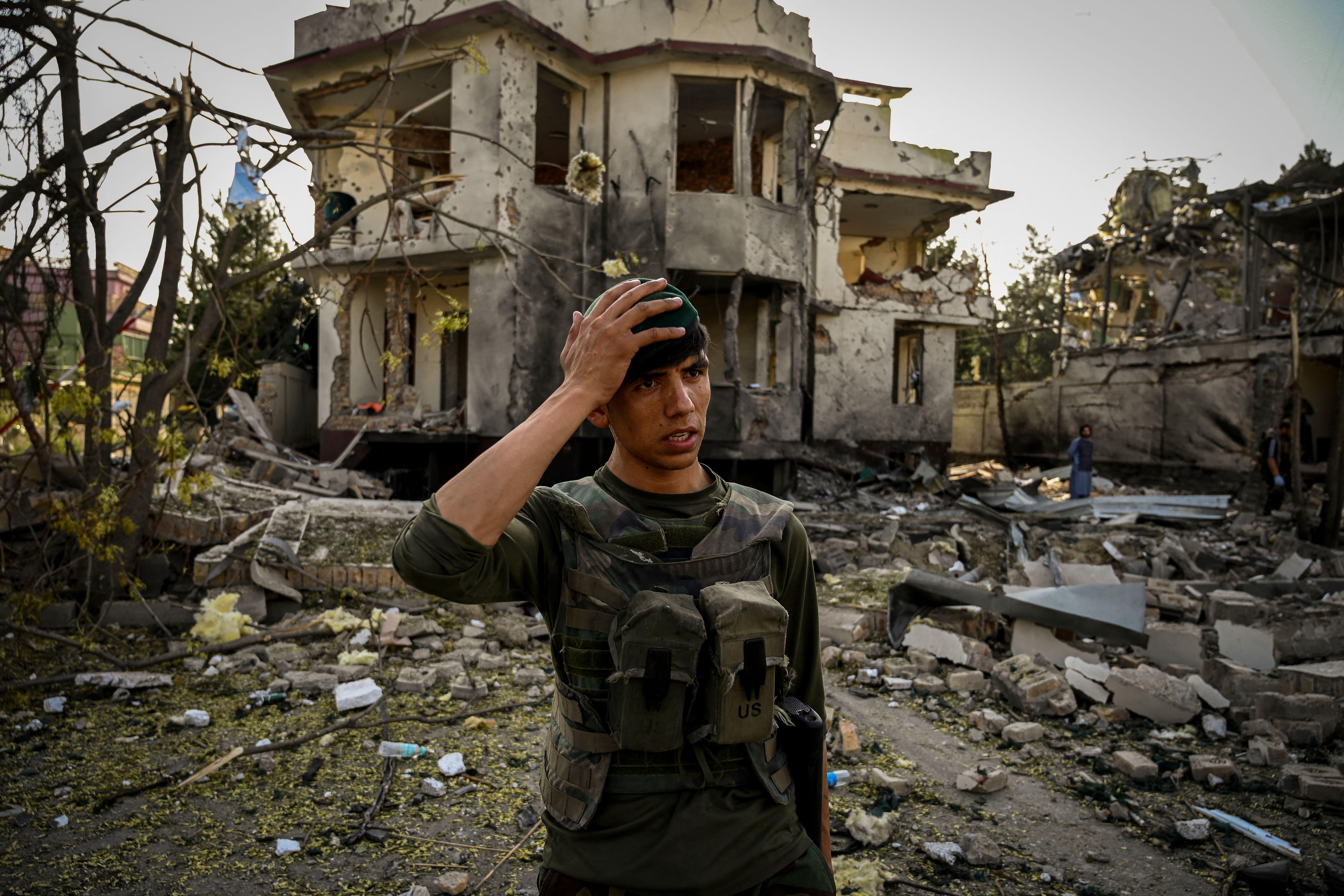 An Afghan man wearing U.S. military gear stands in front of rubble putting his right hand to his head in apparent dismay