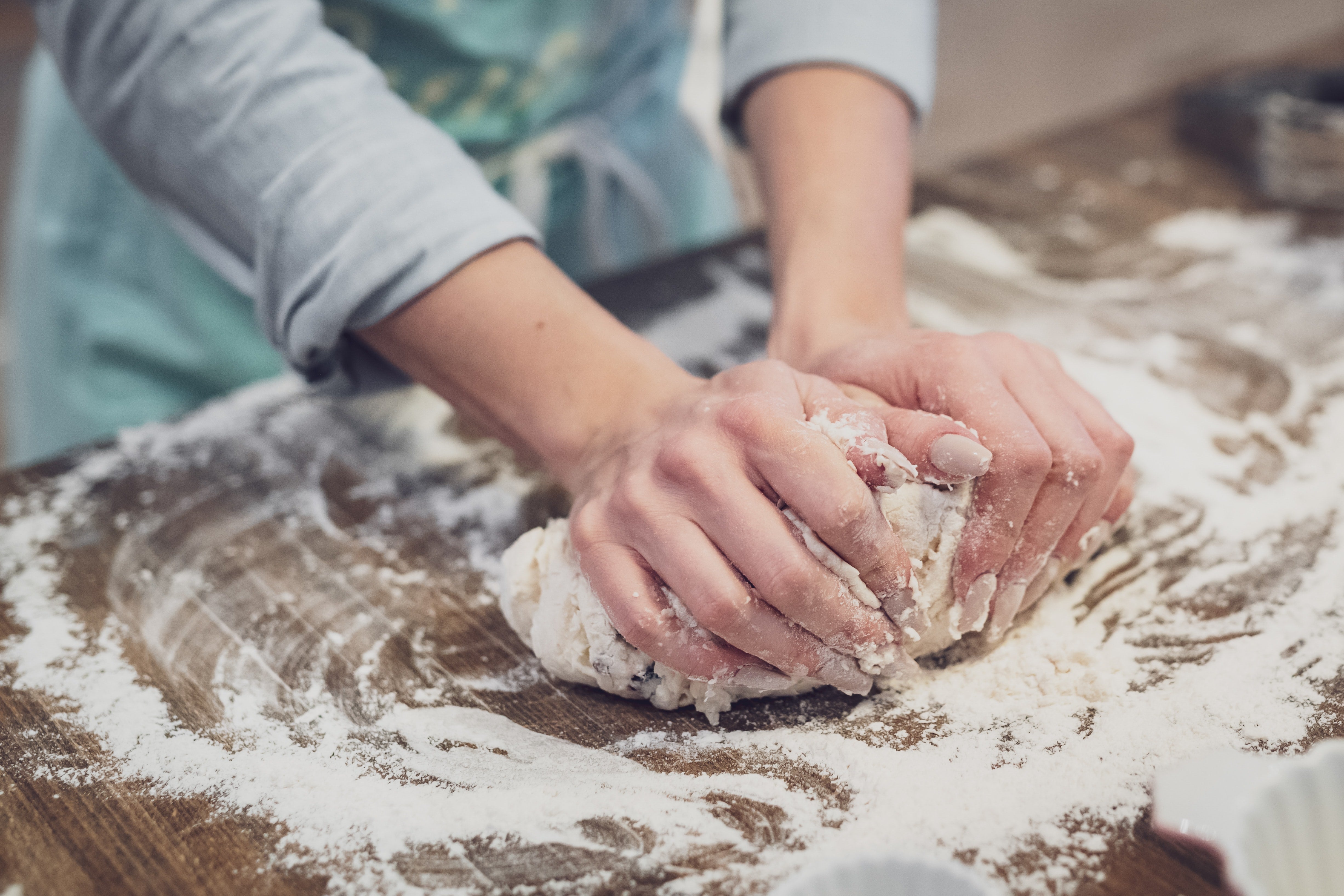A pair of hands kneads a ball of dough on well-floured wooden surface.