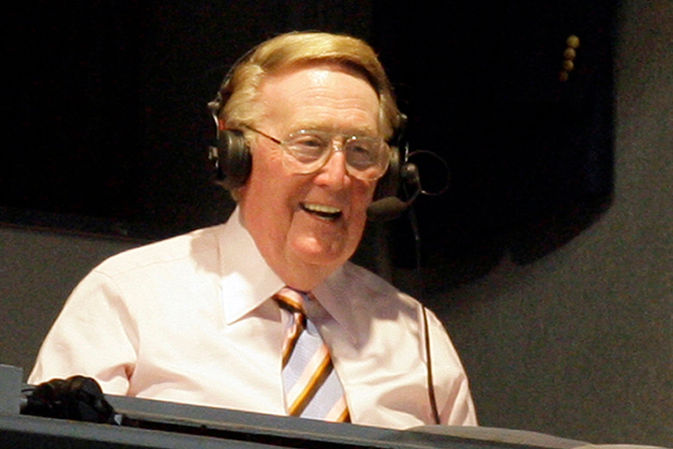 Scully smiling in a headset in the broadcast booth at the stadium.