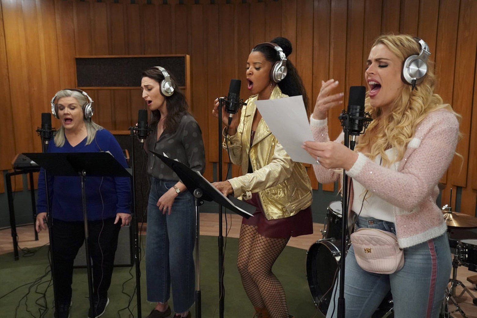 Four women with headphones on sing into microphones in a studio.
