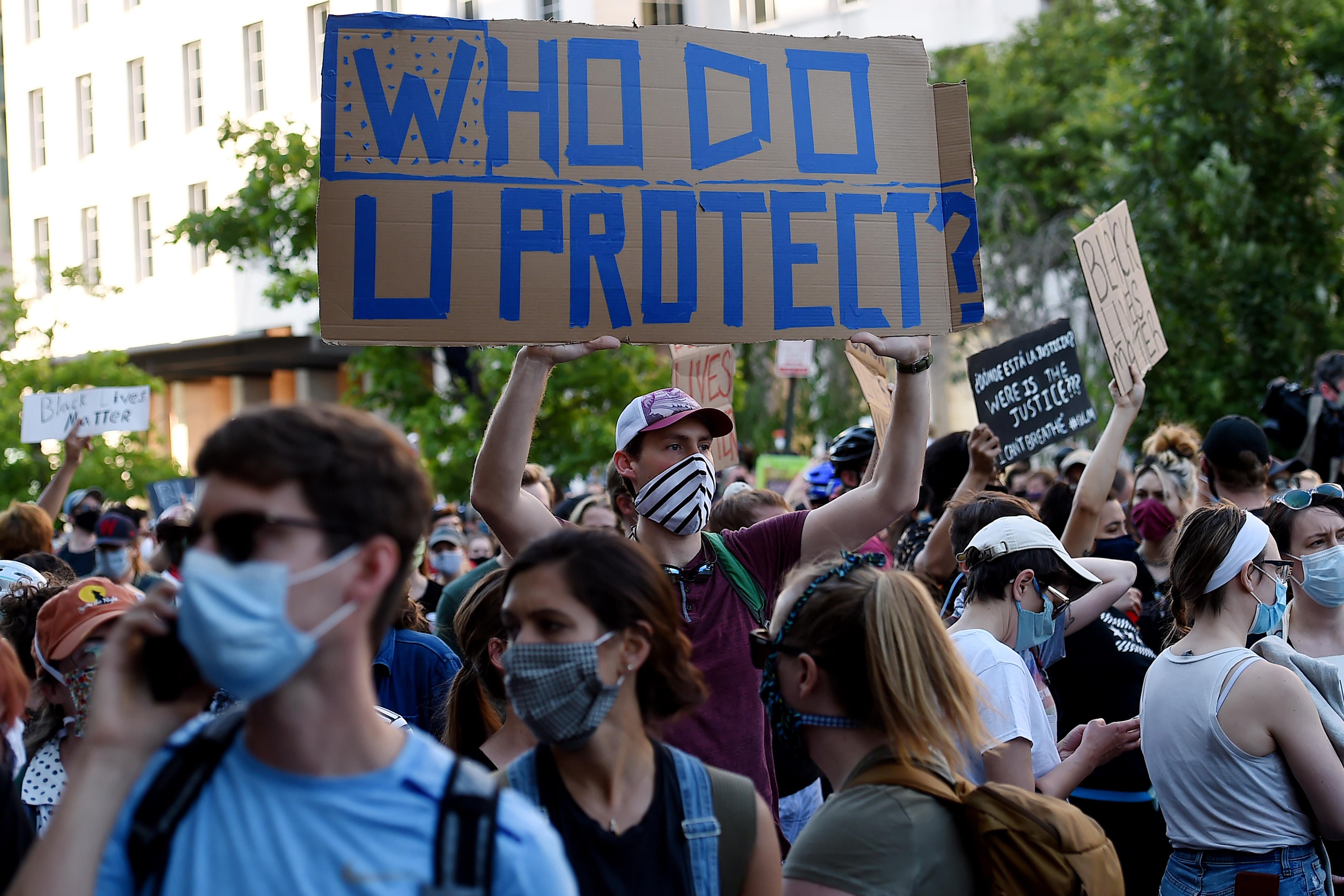 Amid a group of protesters wearing masks, a man holds a sign that says "Who Do U Protect?"