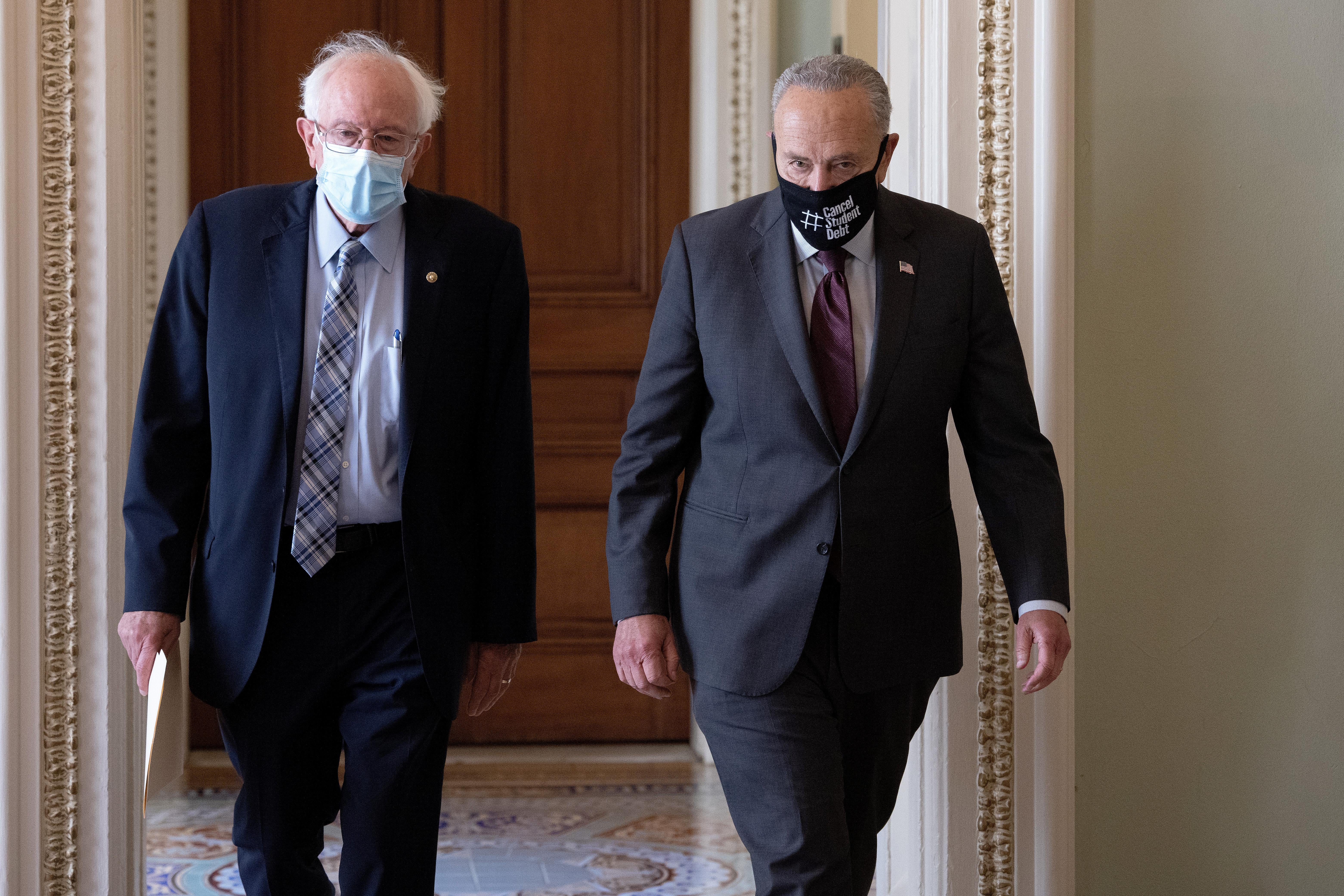 Bernie Sanders and Chuck Schumer walk side by side in a hallway, both wearing masks
