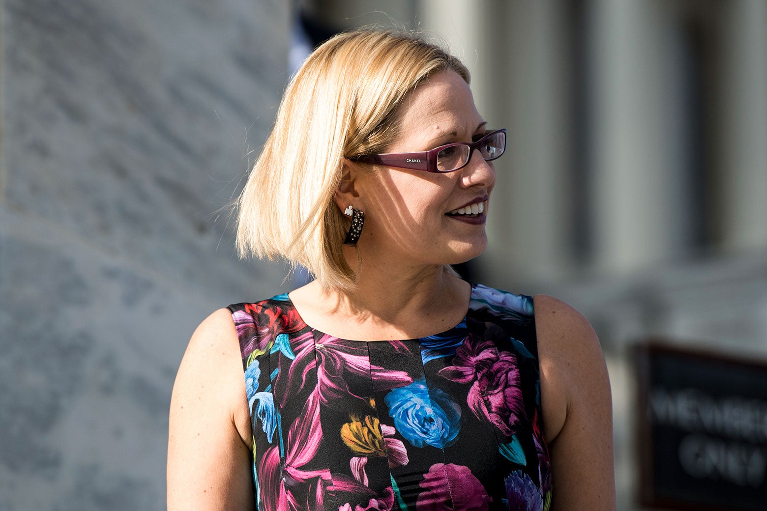 Kyrsten Sinema on the House steps wearing a multicolored floral sleeveless dress, a pair of chunky Chanel glasses, and glittery hoop earrings.