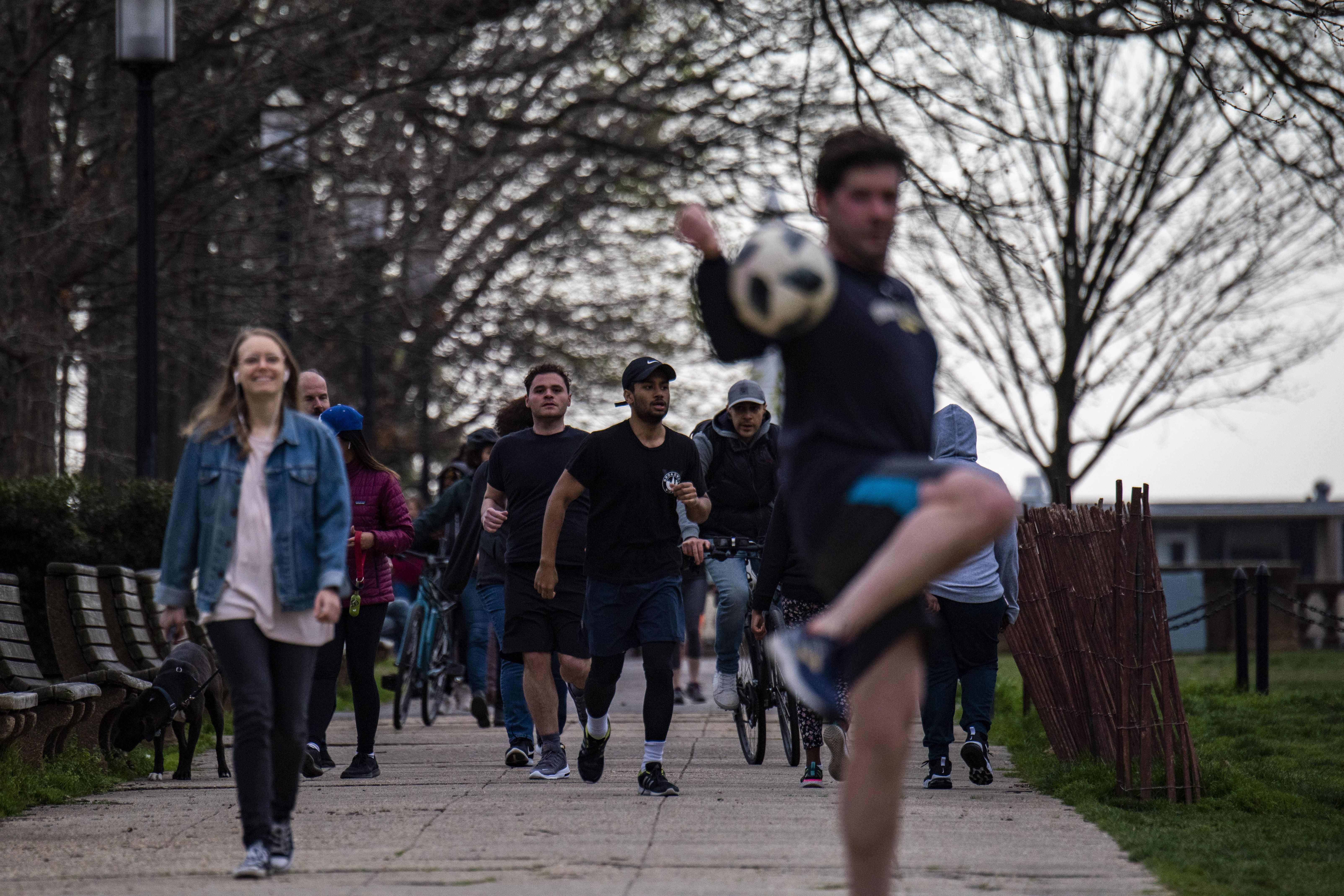 People walk and jog, and a man kicks a soccer ball, on a crowded path.