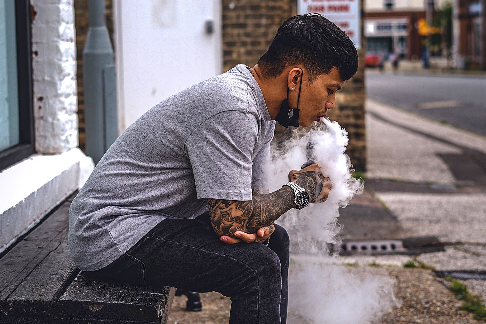A man sitting on a bench hunched over and blowing out a thick cloud of vape smoke.