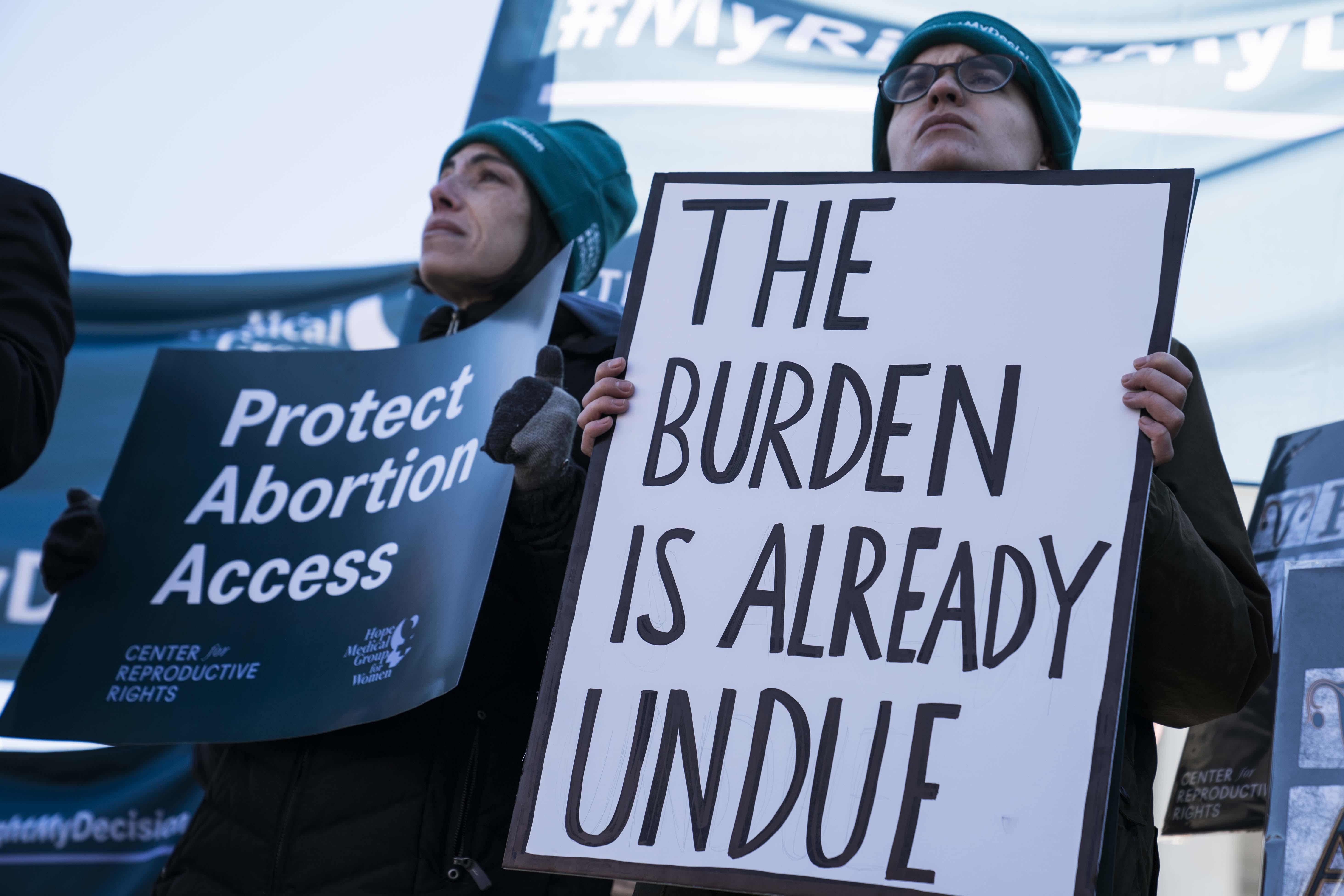 A woman in a beanie holds a sign that says "Protect Abortion Access." A person next to her holds a sign that says "The Burden Is Already Undue."