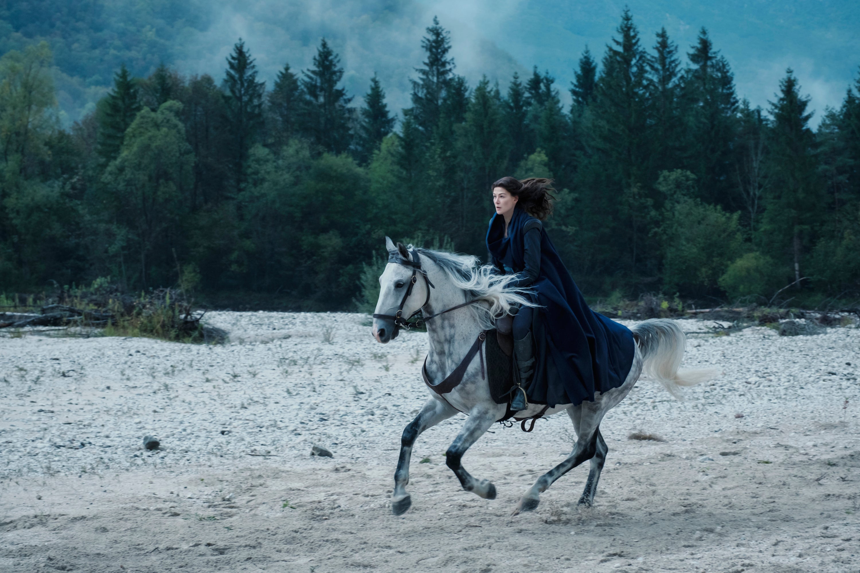 A woman in a cloak rides a white dappled horse in front of a tall forest