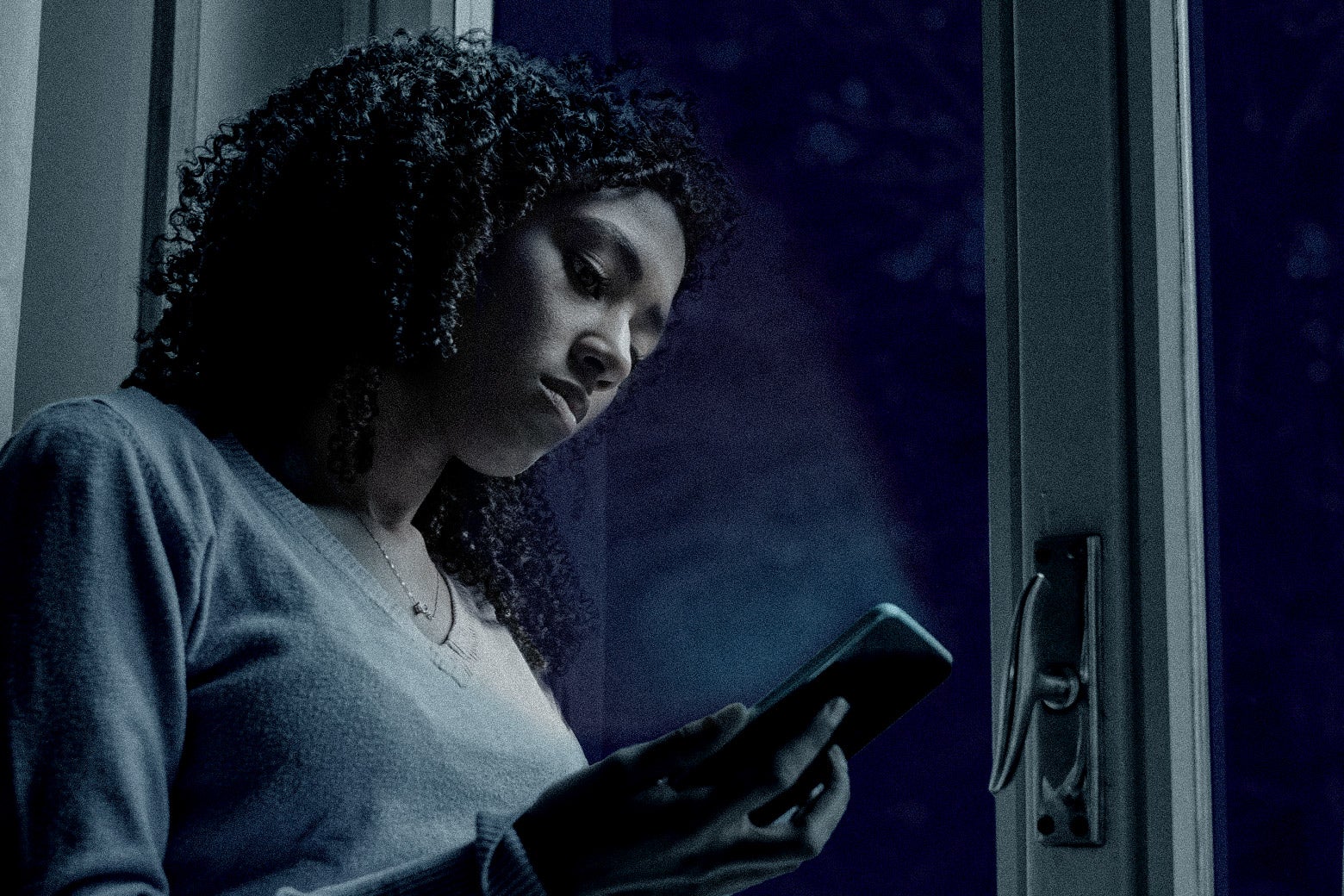 A teenage girl stares at her cell phone, indoors. It's dark outside.