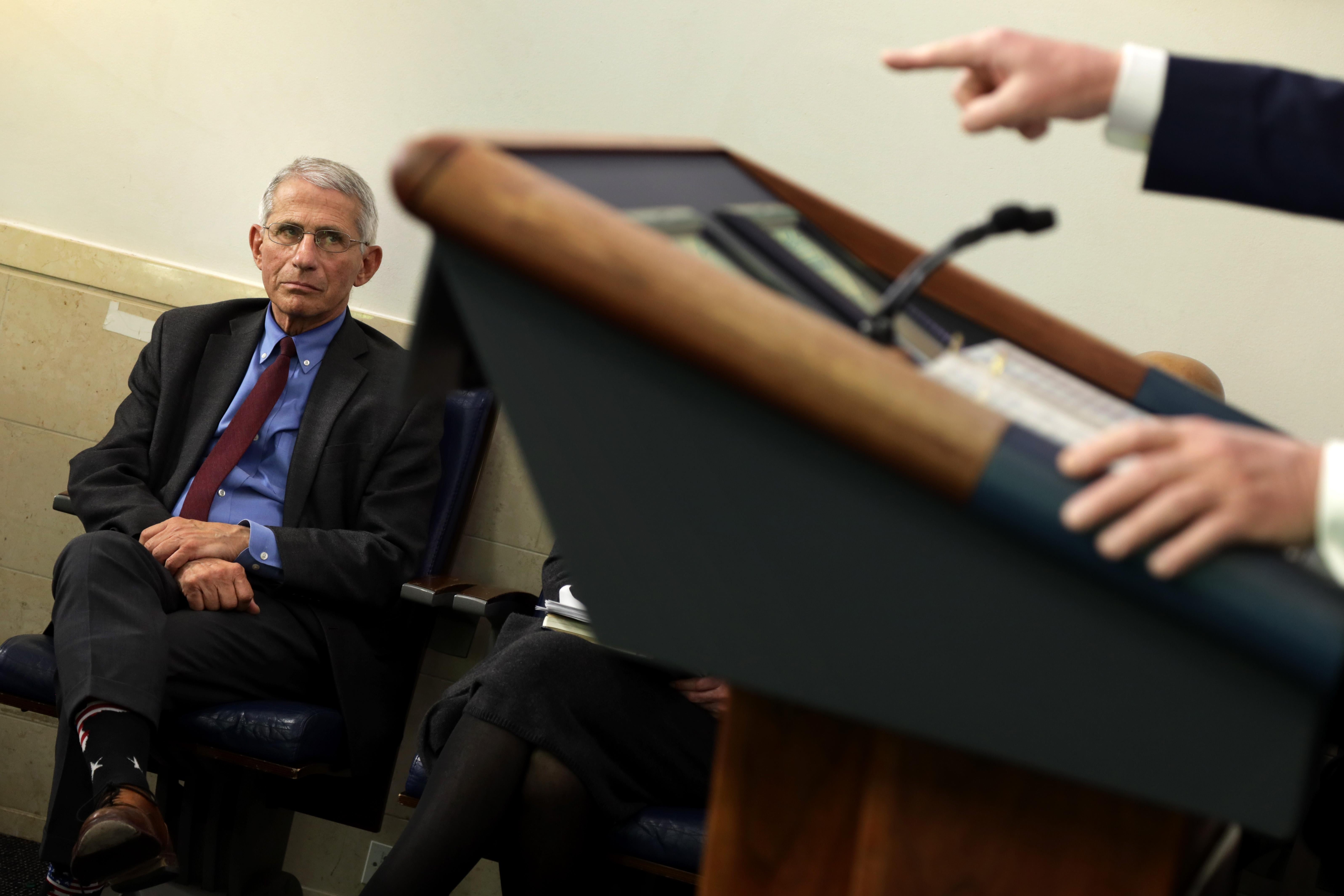 Fauci sits in a chair and watches as Trump stands at the podium, pointing toward the audience