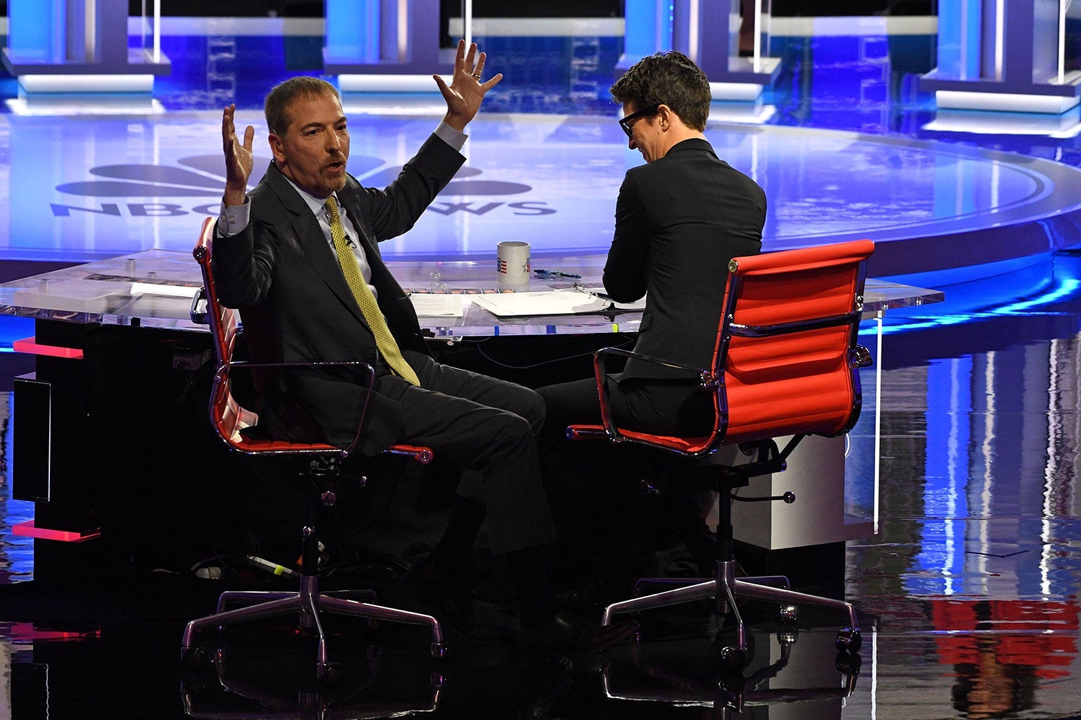 Chuck Todd turns around to face the audience, arms raised in frustration, next to Rachel Maddow. They're both sitting at a desk on the debate stage.