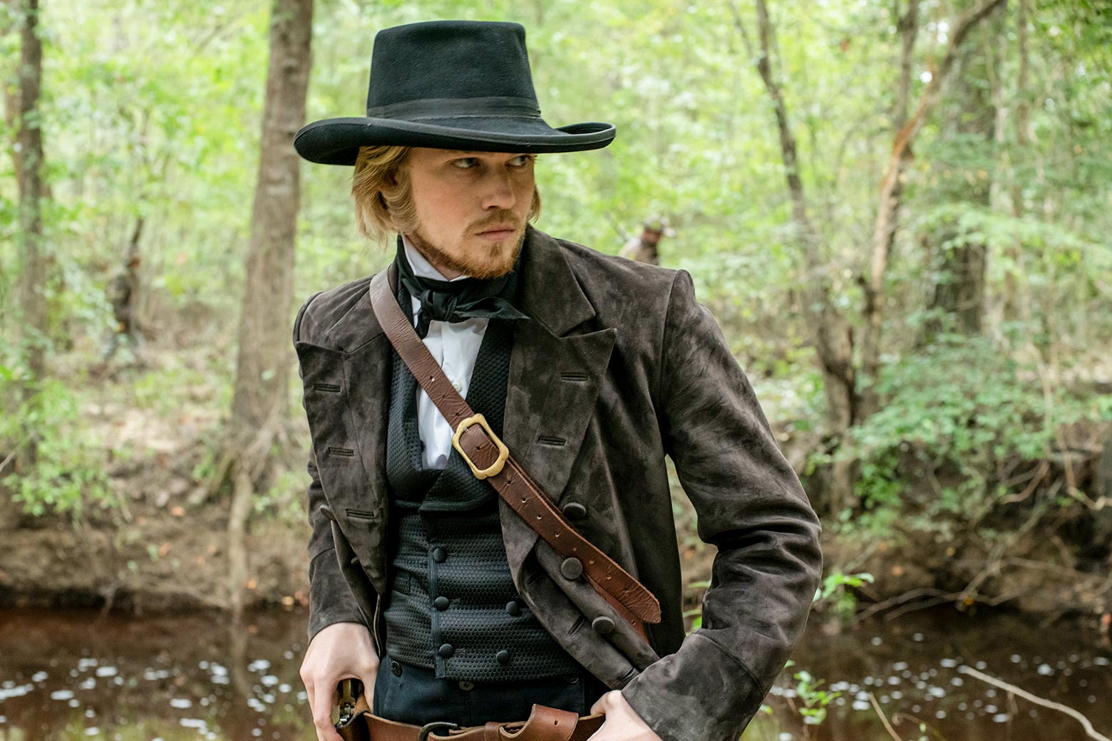 Joe Alwyn as Gideon, wearing 19th century garb and looking around in a forest.