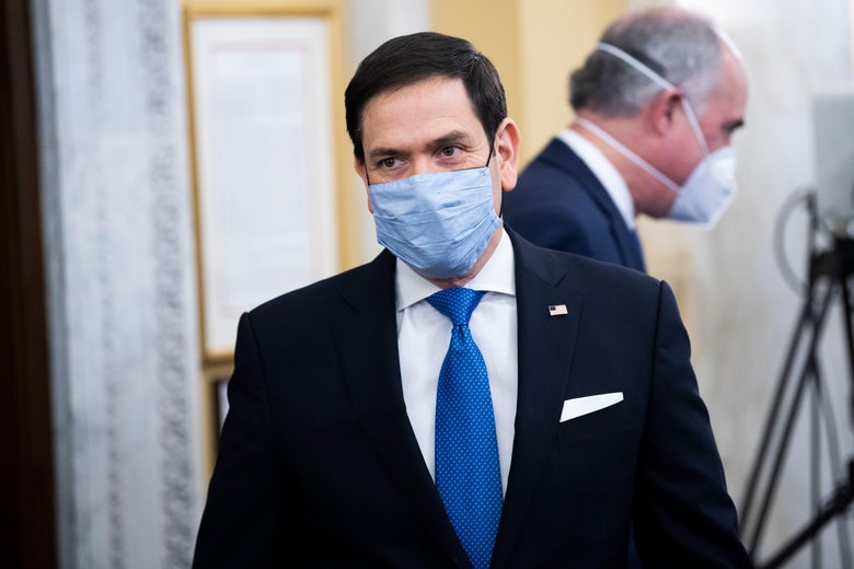 Marco Rubio in a suit and mask walking toward the camera.