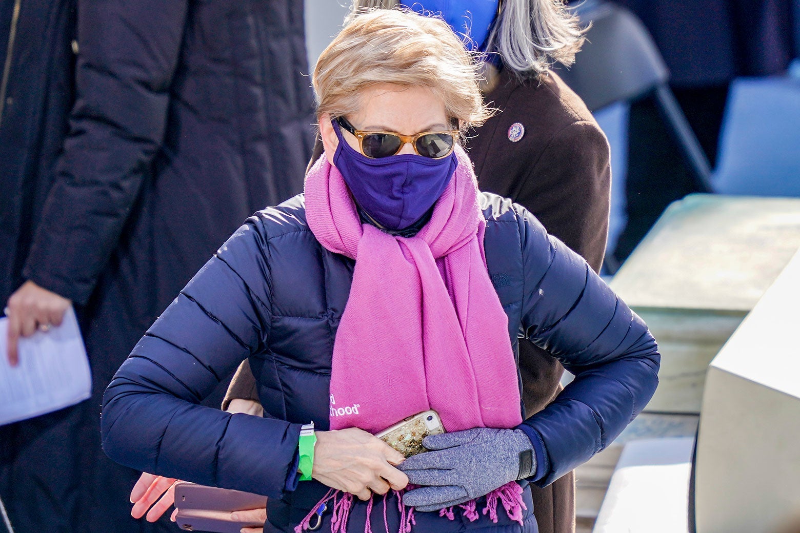 Elizabeth Warren wears sunglasses, a purple face mask, a pink scarf, and a purple puffer coat.