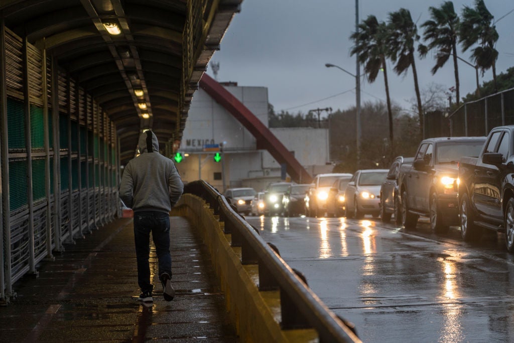 A man in a hoodie walks on a sidewalk as a line of cars pass him against a rainy, gray sky.