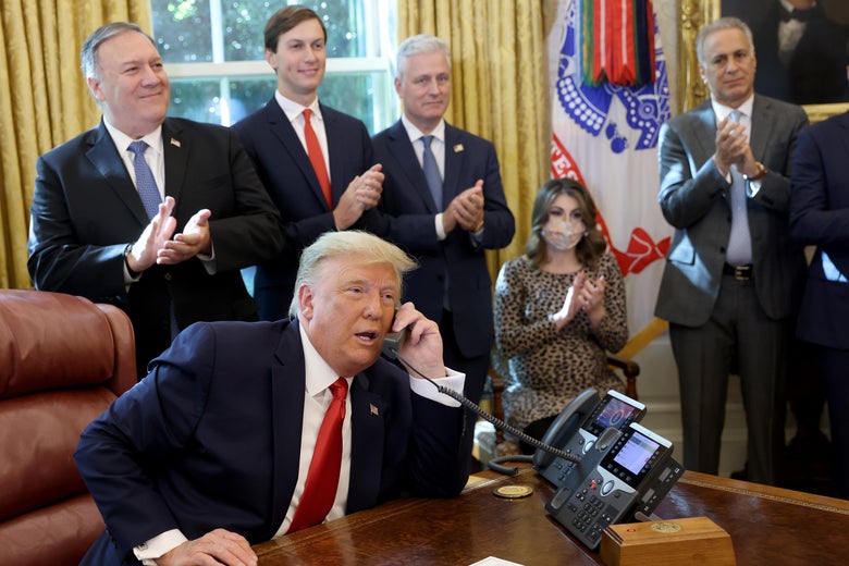 Donald Trump talks on a phone behind the Resolute desk while staffers, including Mike Pompeo and Jared Kushner, applaud.