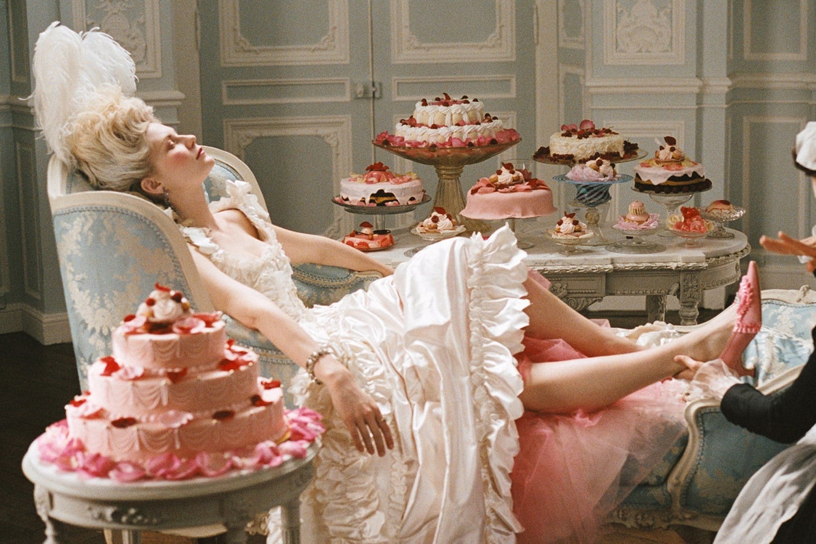 A maid holds Kirsten Dunst's foot who is dressed as Marie Antoinette in an elaborate white dress and headdress while lounging surrounded by cakes and pastries. 