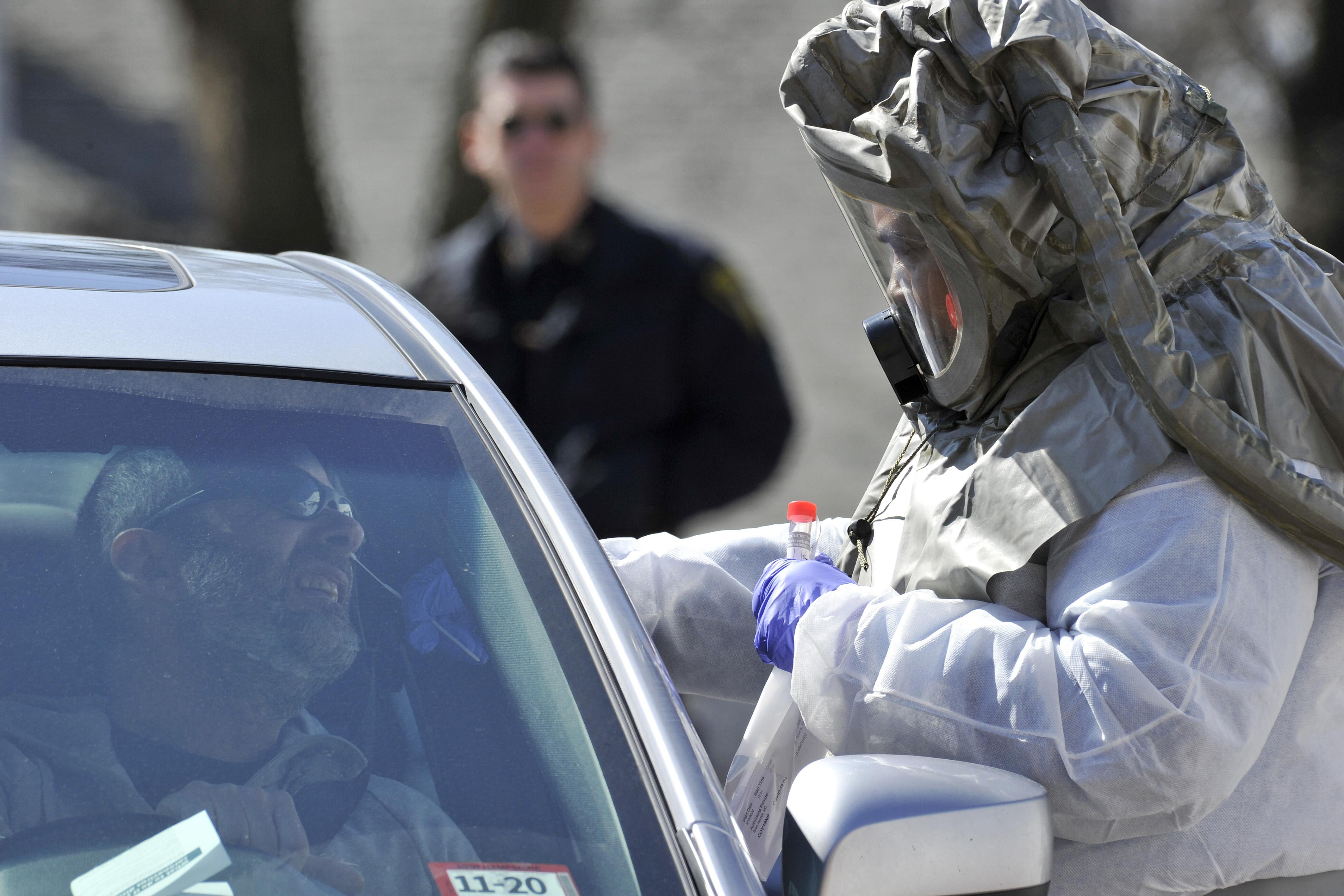 A man sits in his car with his face angled up as a person in protective gear takes a swab of his nose.
