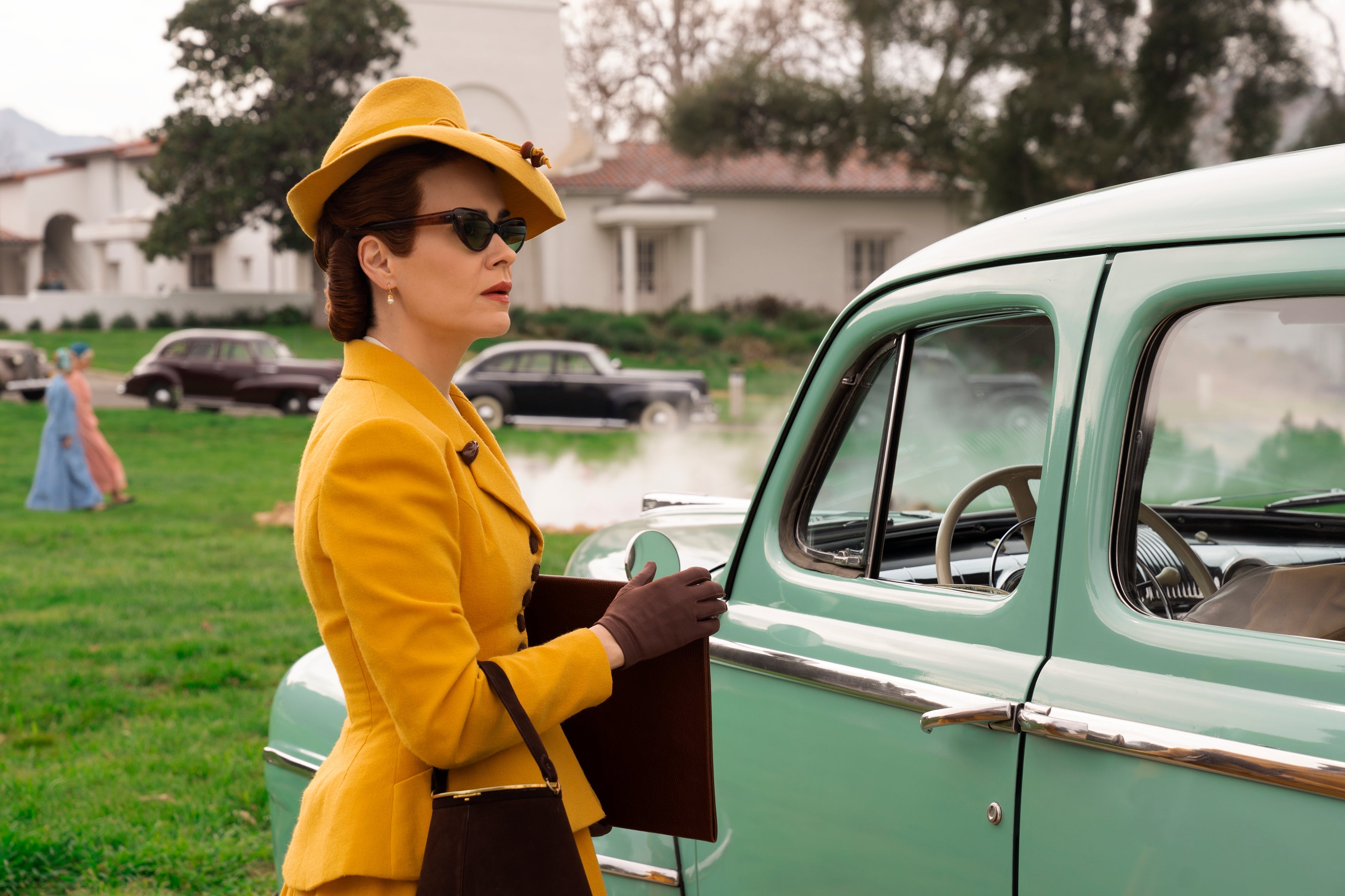 Sarah Paulson as Mildred Ratched, in a yellow suit with sunglasses in front of an old-fashioned automobile.