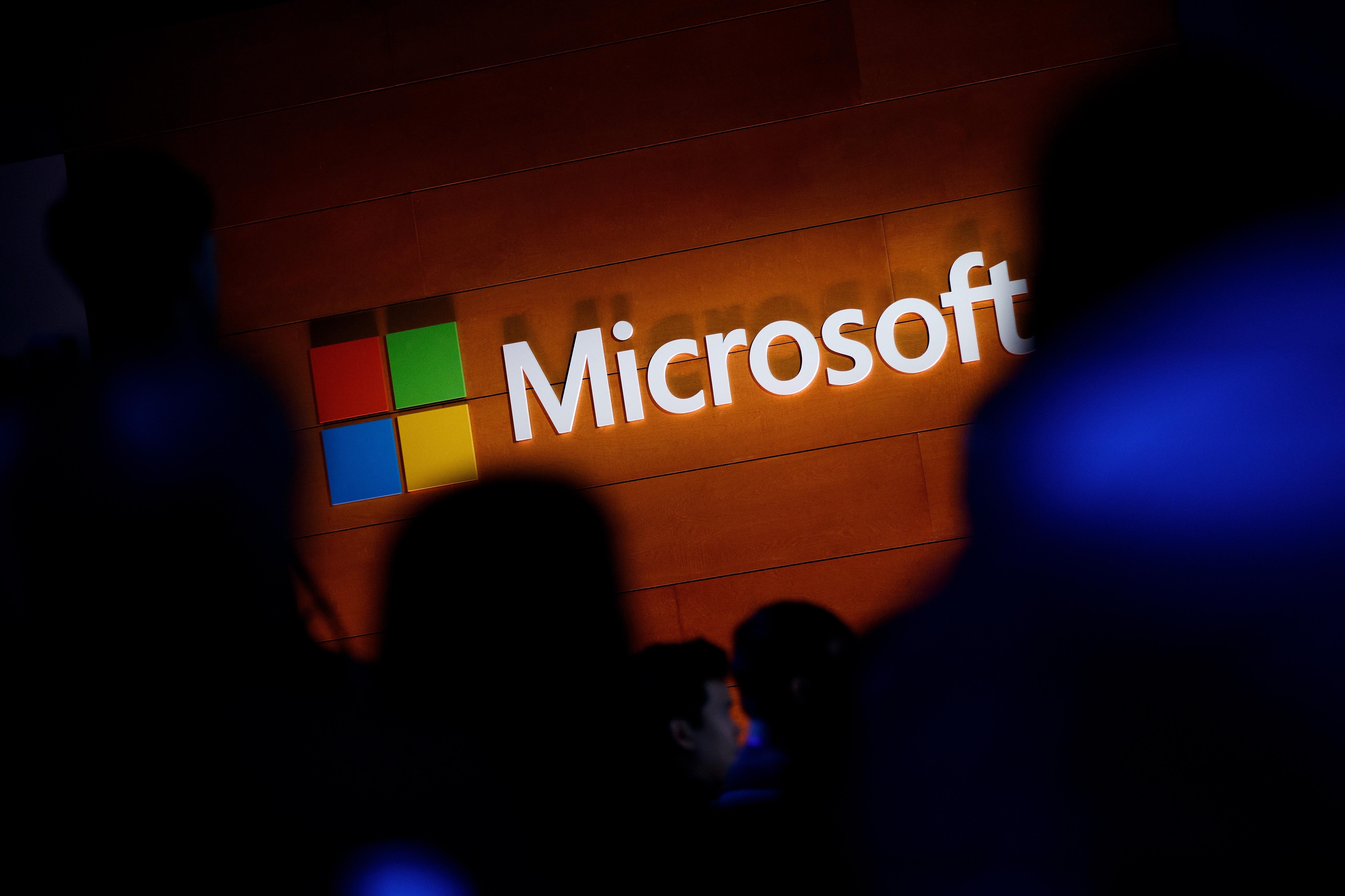 The Microsoft logo is illuminated on a wooden wall. It is surrounded by a crowd of dark silhouettes.