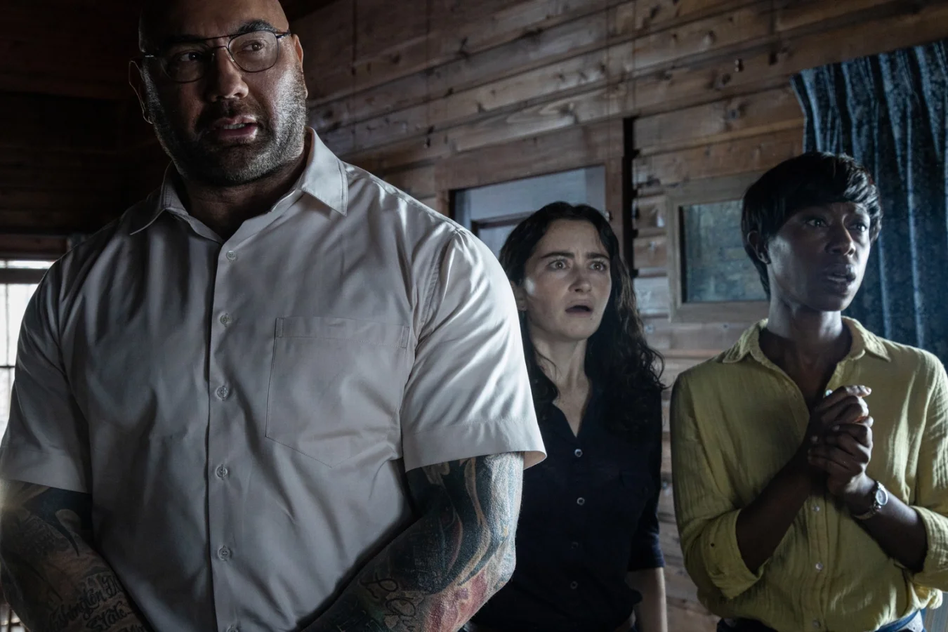 A large tattooed man and two smaller women stand in a wooden cabin, their faces in shock.
