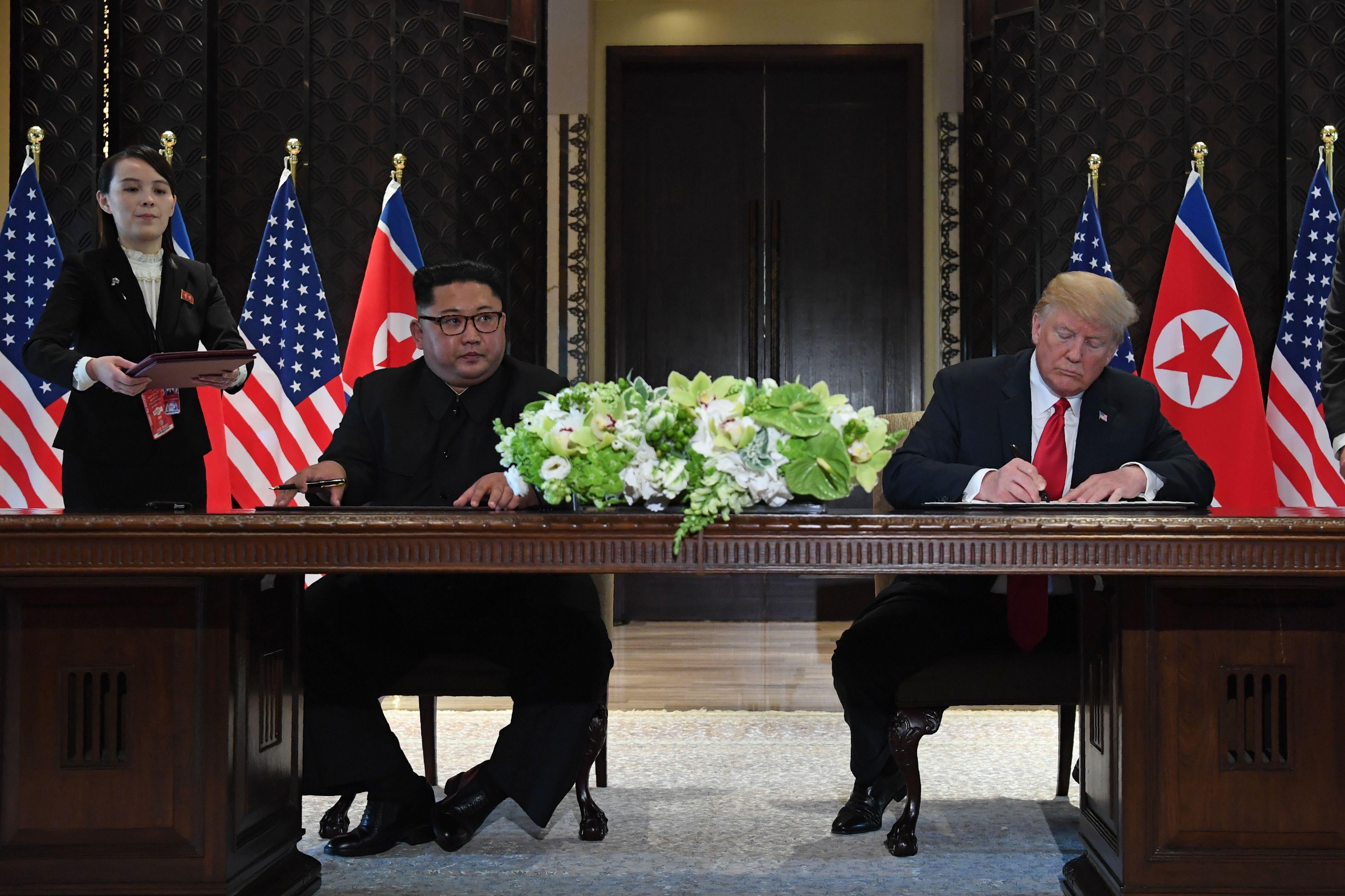 Kim Jong-un and Donald Trump sit at a table and sign the joint statement, with Mike Pompeo standing over Trump.