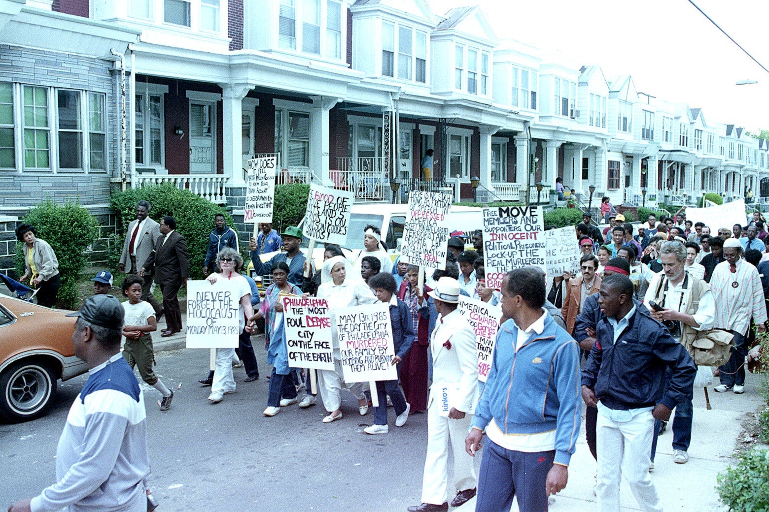 People march in the road past row houses holding signs in support of MOVE