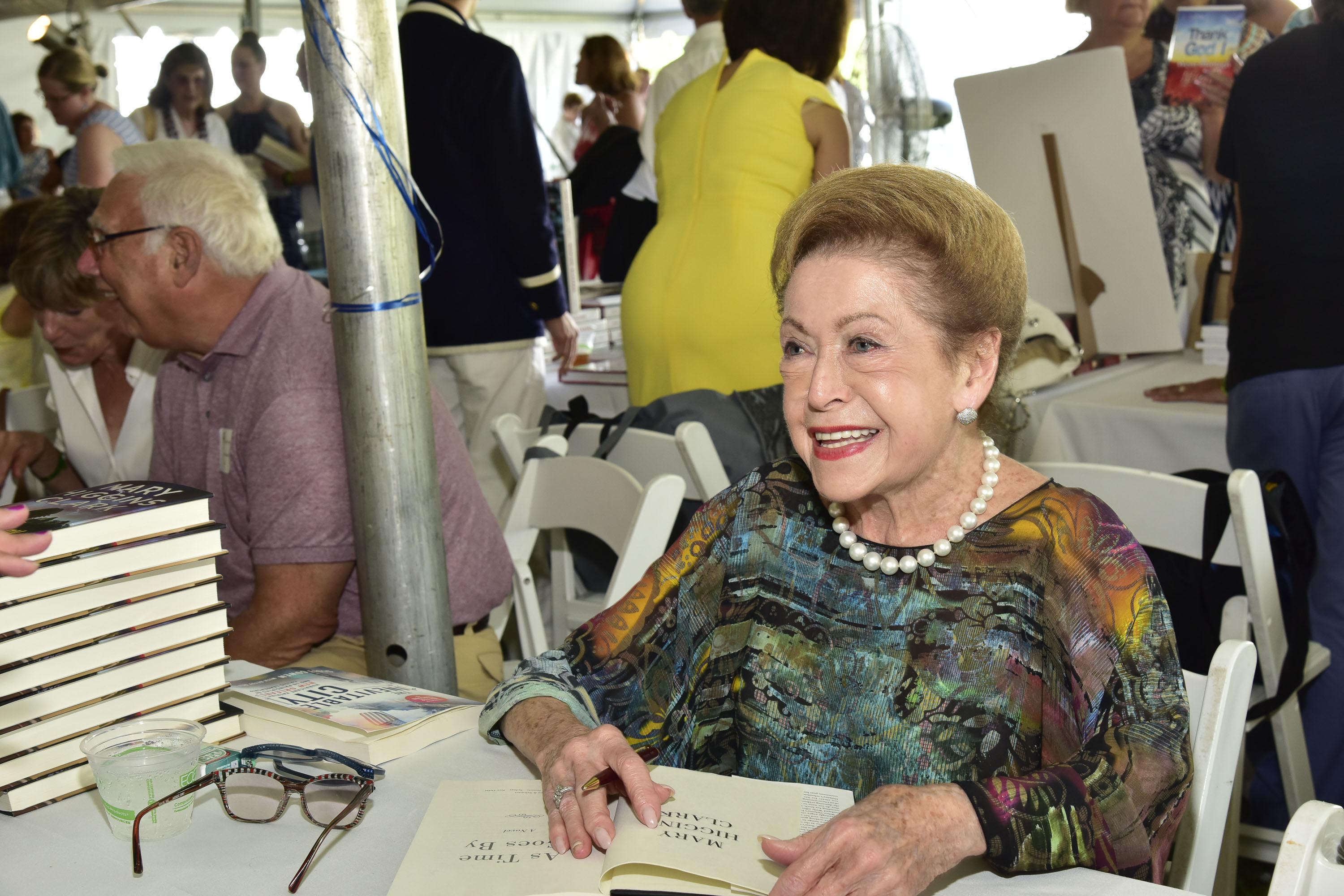 Mary Higgins Clark sits at a folding table with a stack of her books on it. She has one book open to sign, and is smiling.