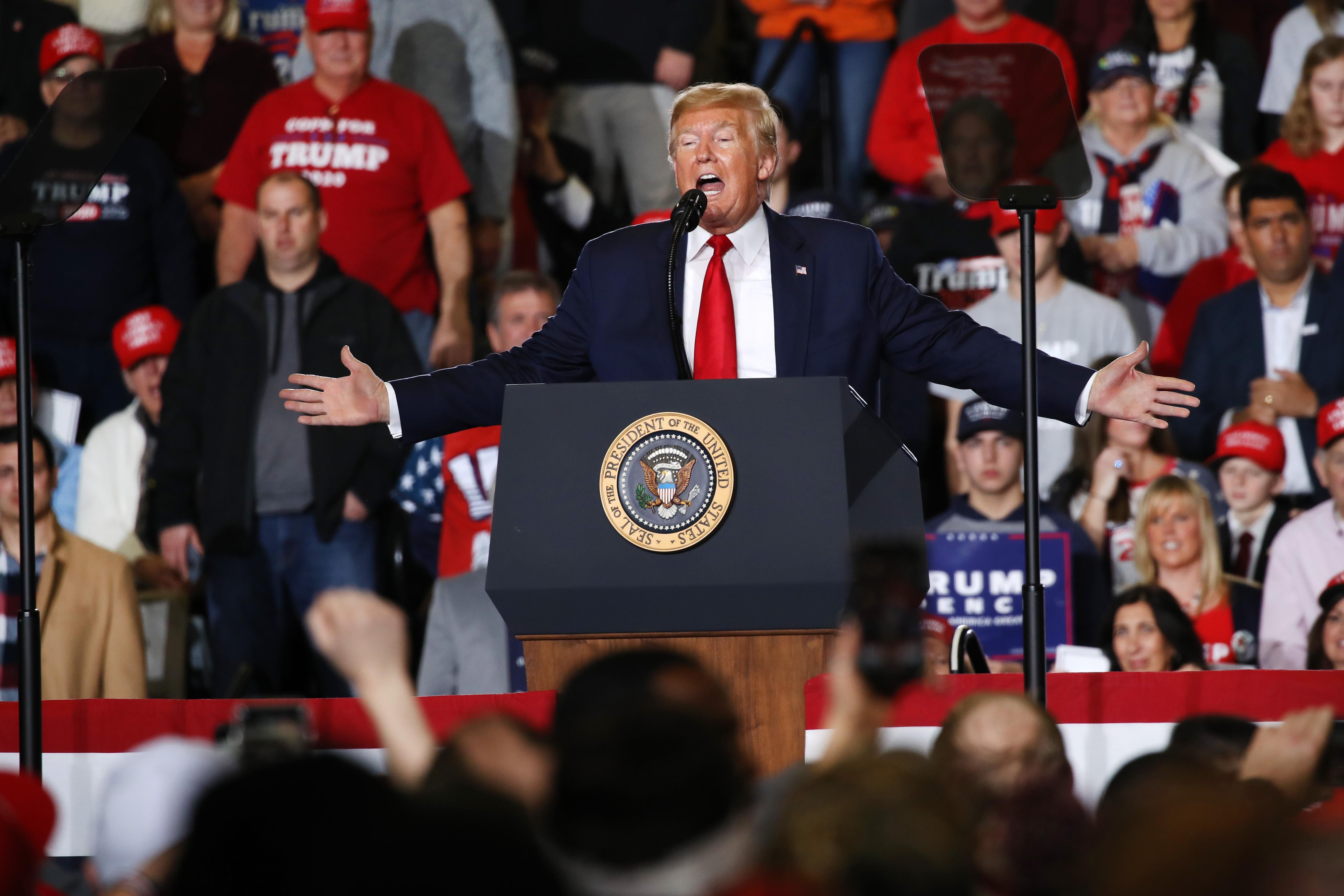 Trump speaks at a podium with his arms out wide and a crowd behind him.