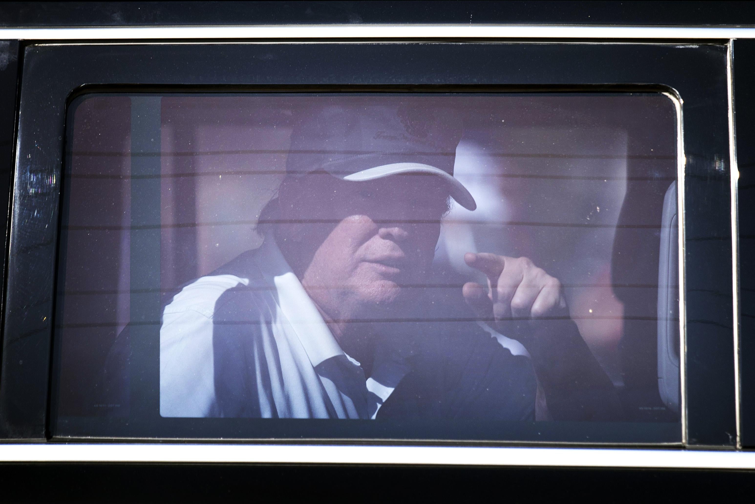 Trump, wearing a white hat, points from inside a limo.
