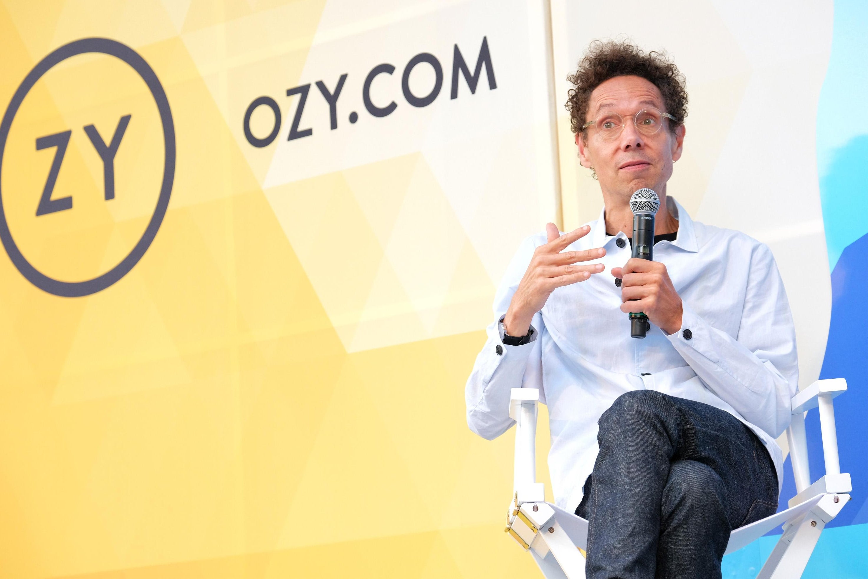 Malcolm Gladwell gesticulates and holds a mic in front of an Ozy-branded backdrop.
