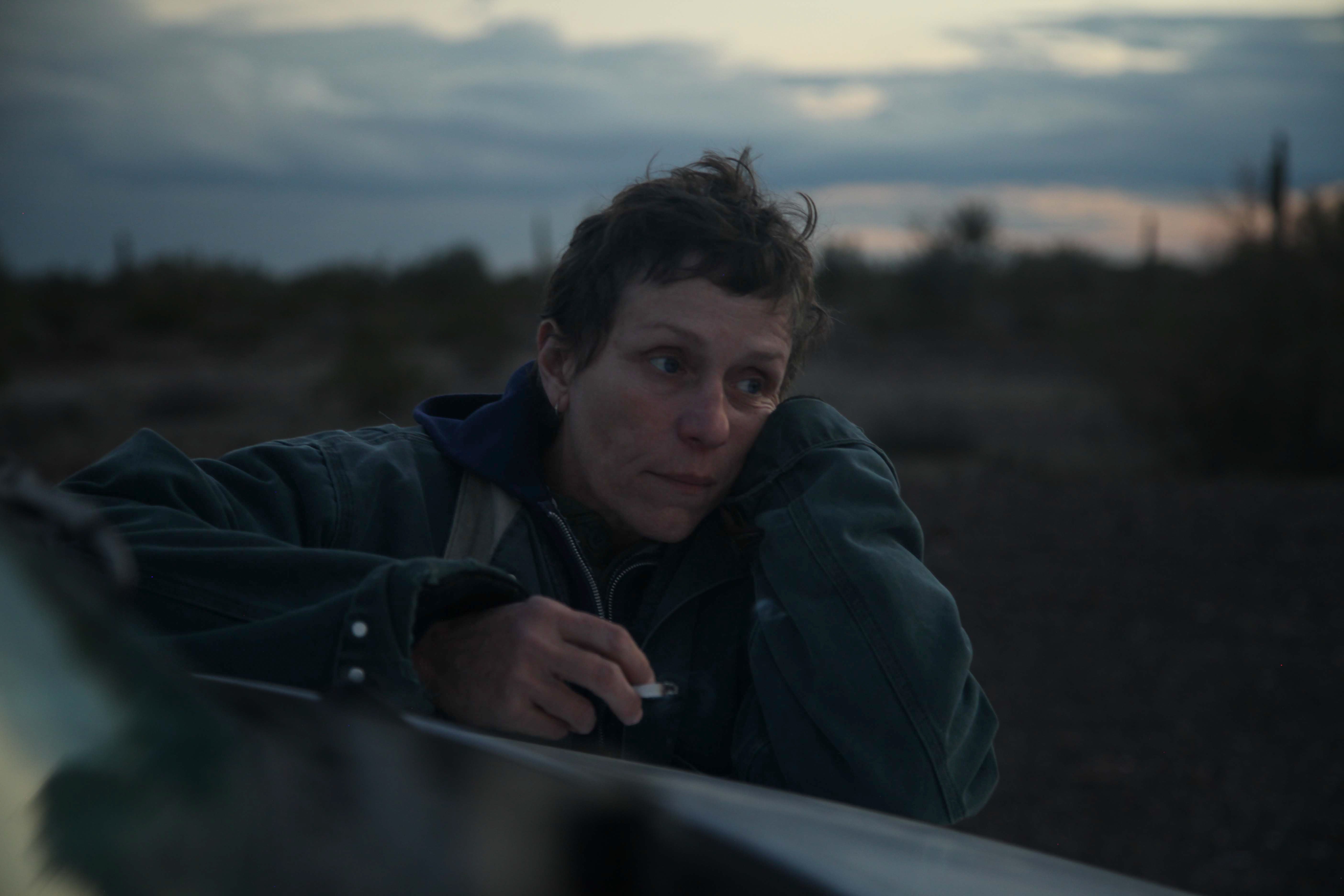 Frances McDormand in short hair, leaning on the hood of a van, a cigarette in one hand
