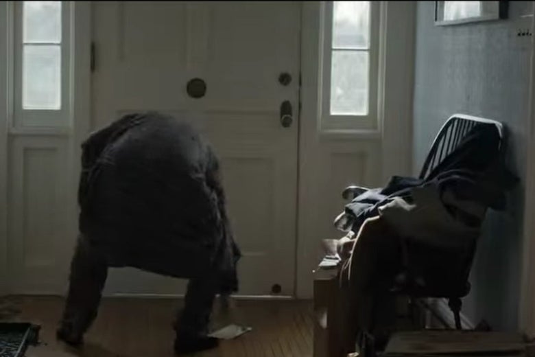 Bill bends down to pick something up in front of his front door, next to a chair piled with clothes and other stuff.