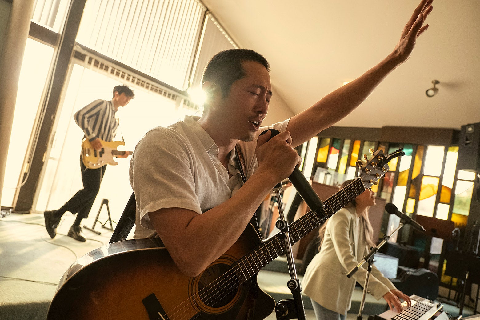 Steven Yuen playing a guitar and singing into a microphone with his left hand in the air.