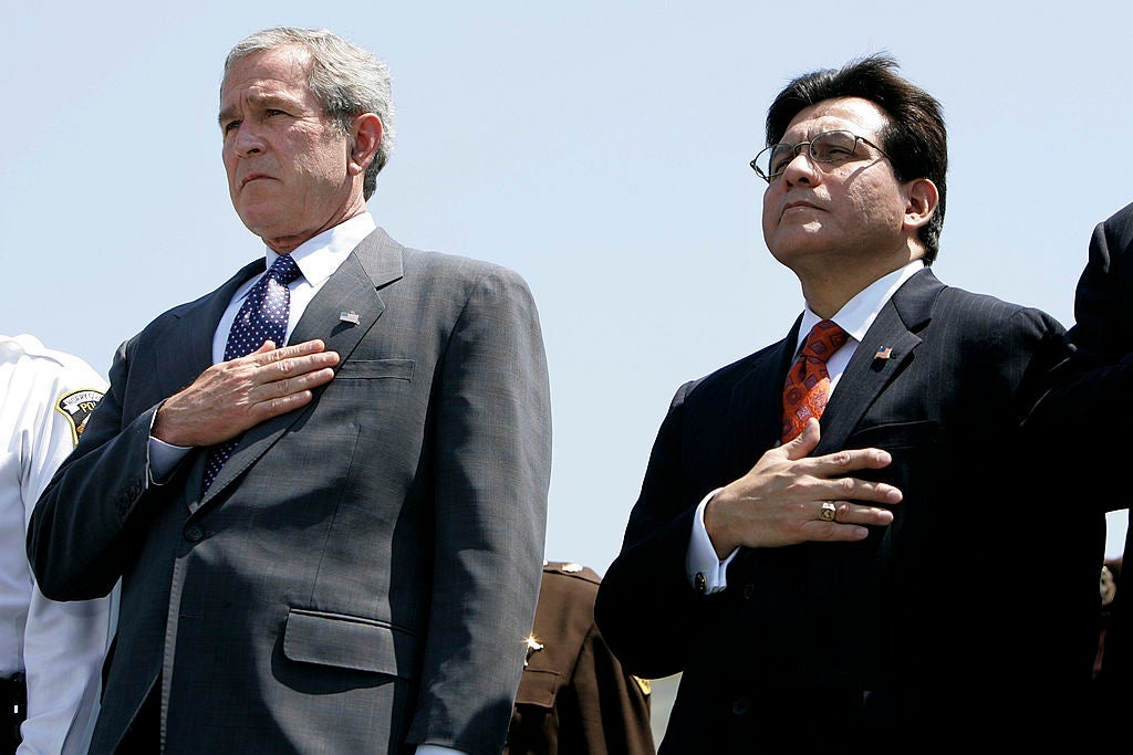 George W. Bush and Alberto Gonzales stand side by side against a blue sky with their hands over their hearts.