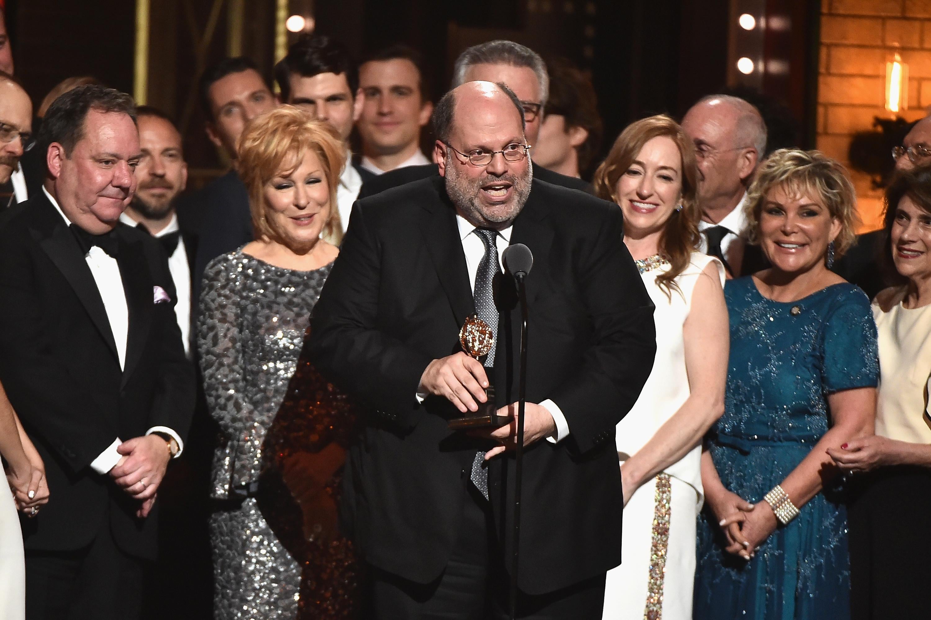 Scott Rudin, in a tuxedo, stands on stage holding a Tony, surrounded by the cast of Hello Dolly!