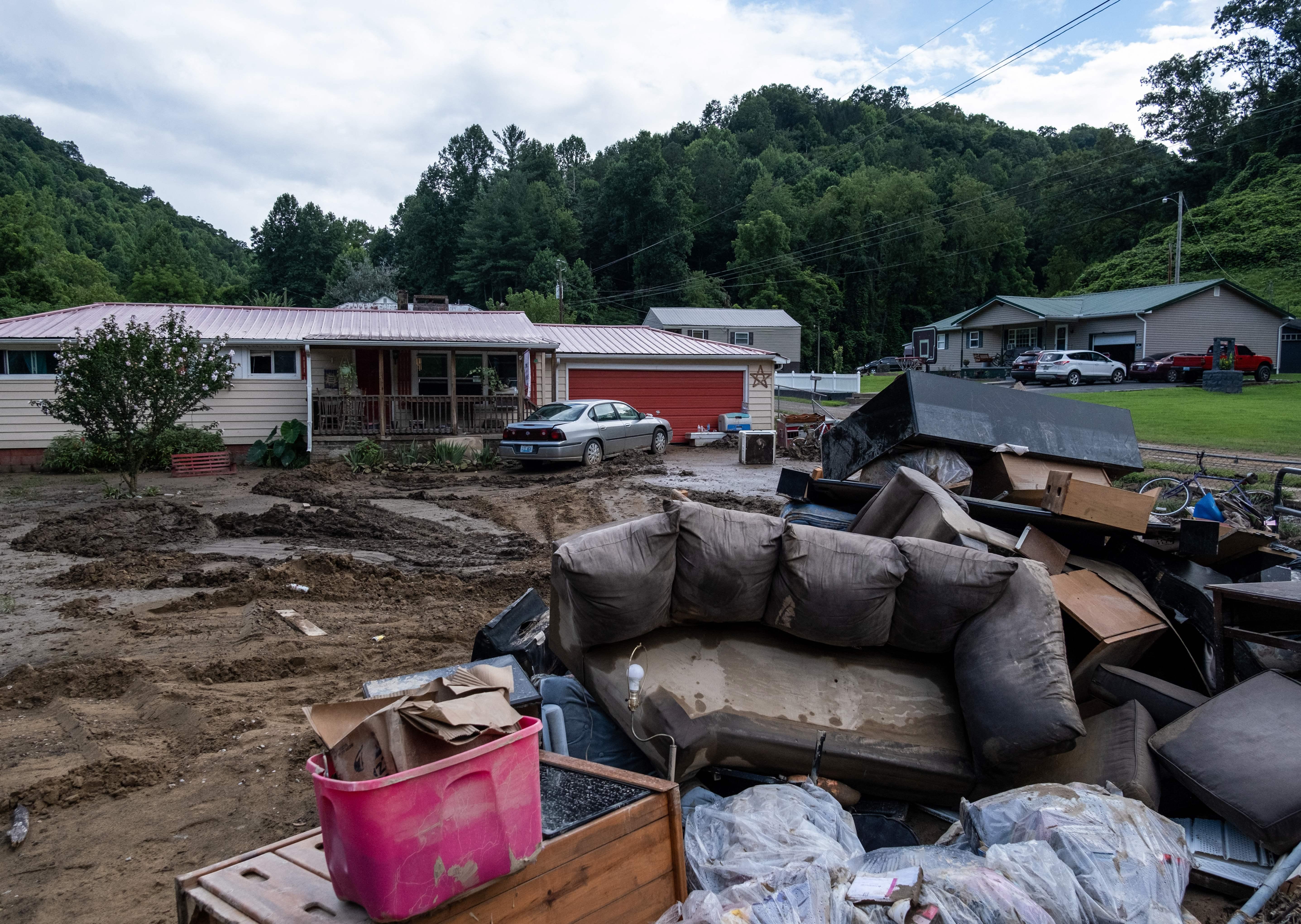 Water-damaged furniture sit in a pile outside of a one-story home surrounded by mud. 