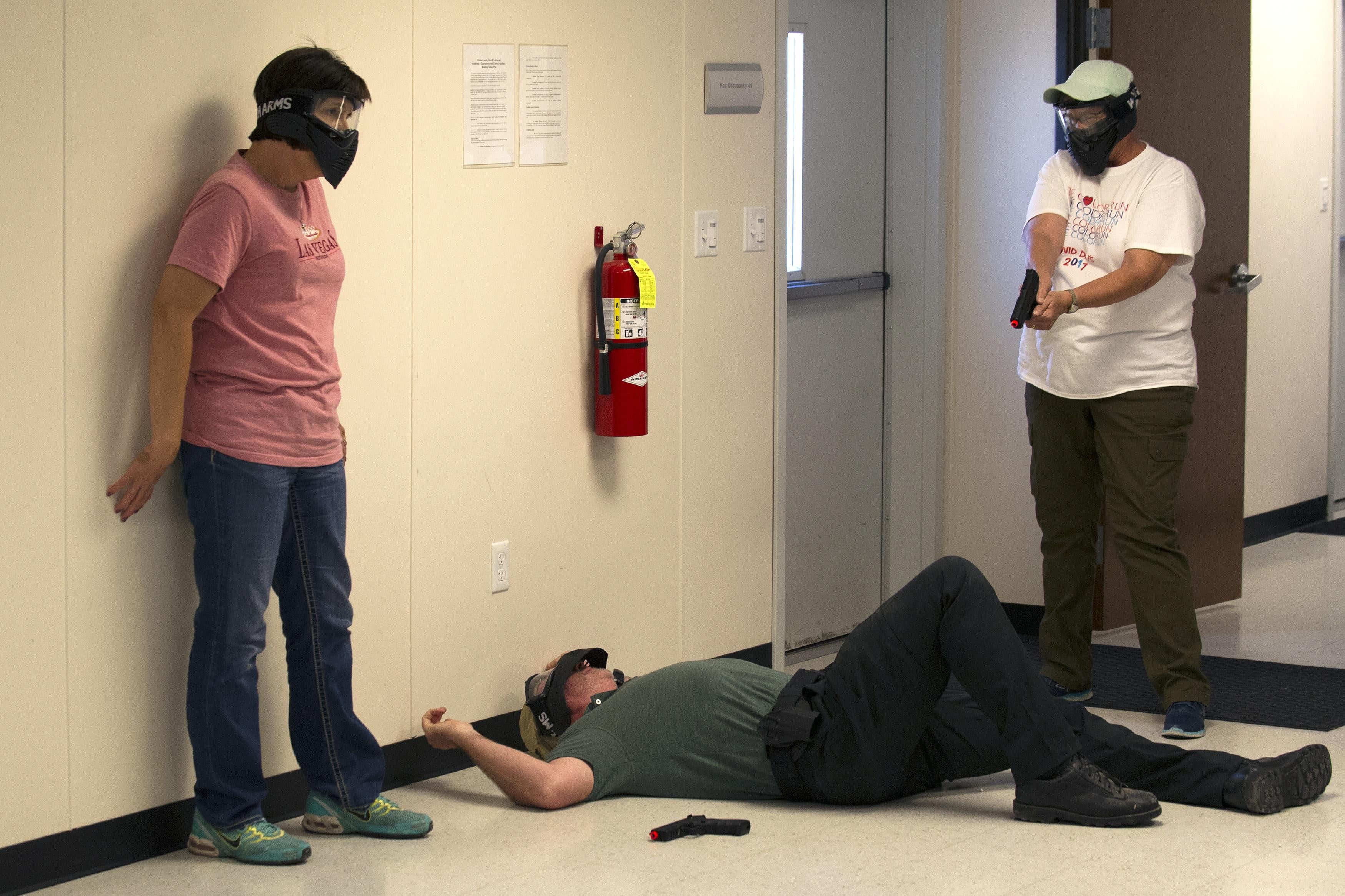 A person wearing a paintball mask points a pellet gun at a man lying on the ground in a hallway. Another person stands next to that man.