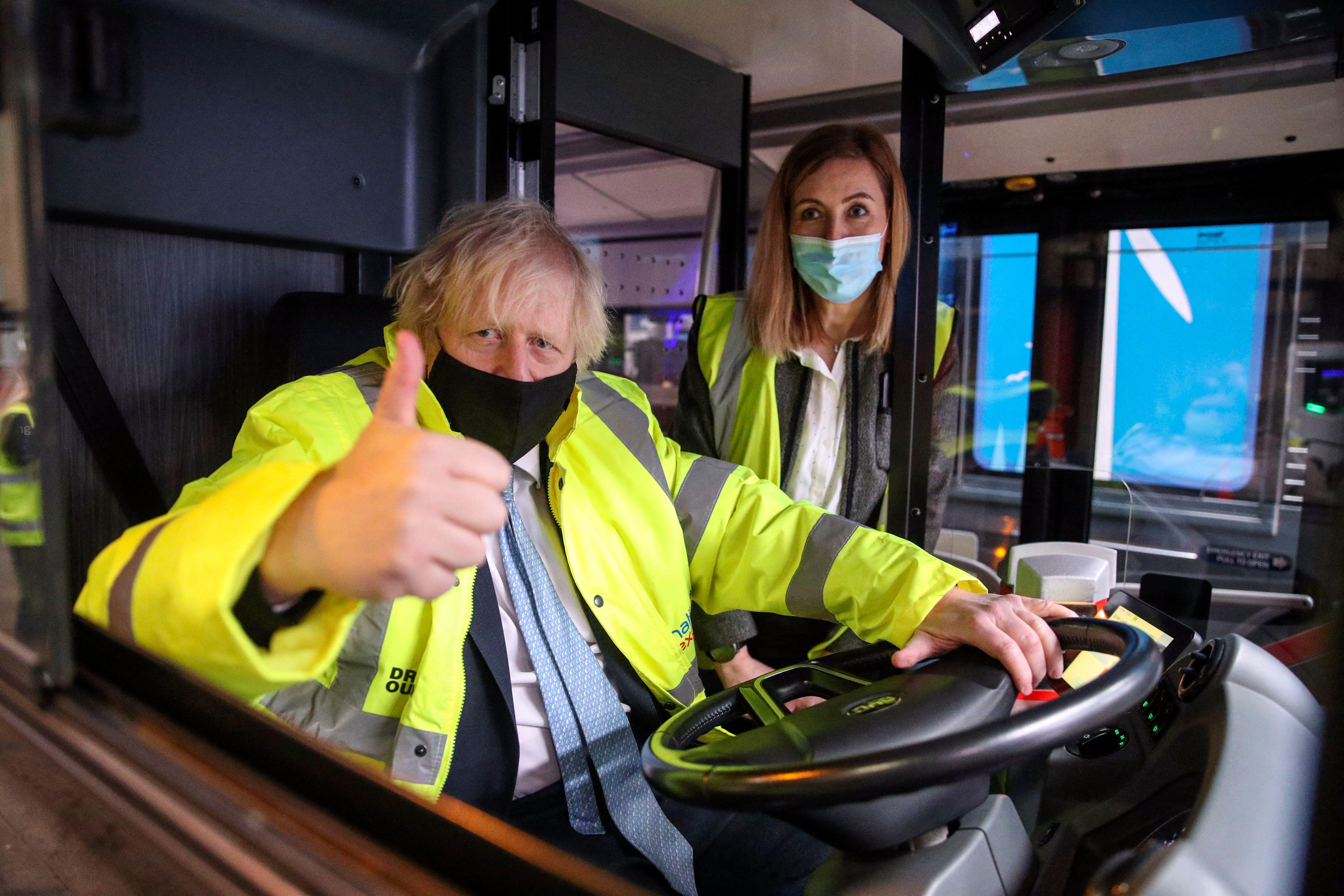 Boris Johnson wears a yellow high-visibility jacket and gives a thumbs-up as he sits behind the wheel of a bus.