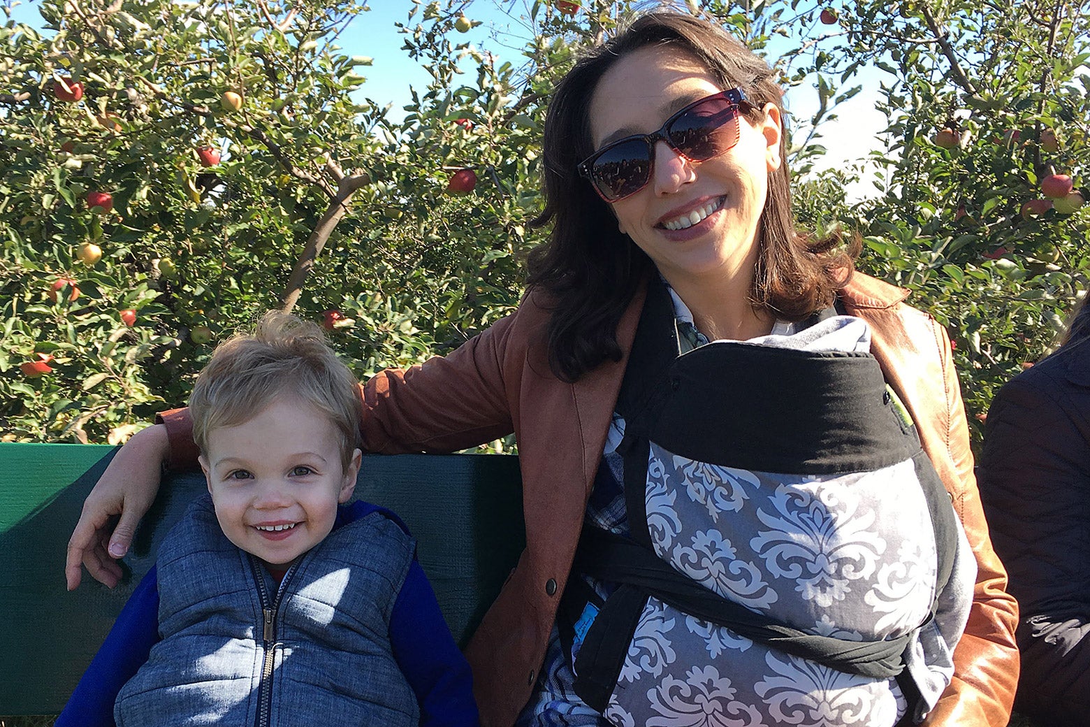 A woman sits next to a small child in an orchard. Another child is strapped to her chest.