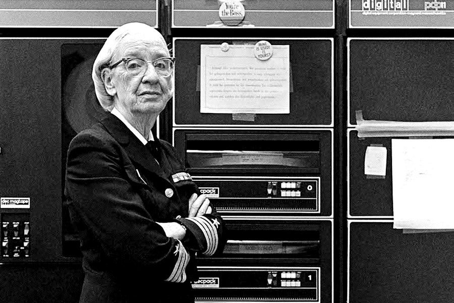 Grace Hopper stands in her Navy uniform, arms crossed, in front of a massive computer.