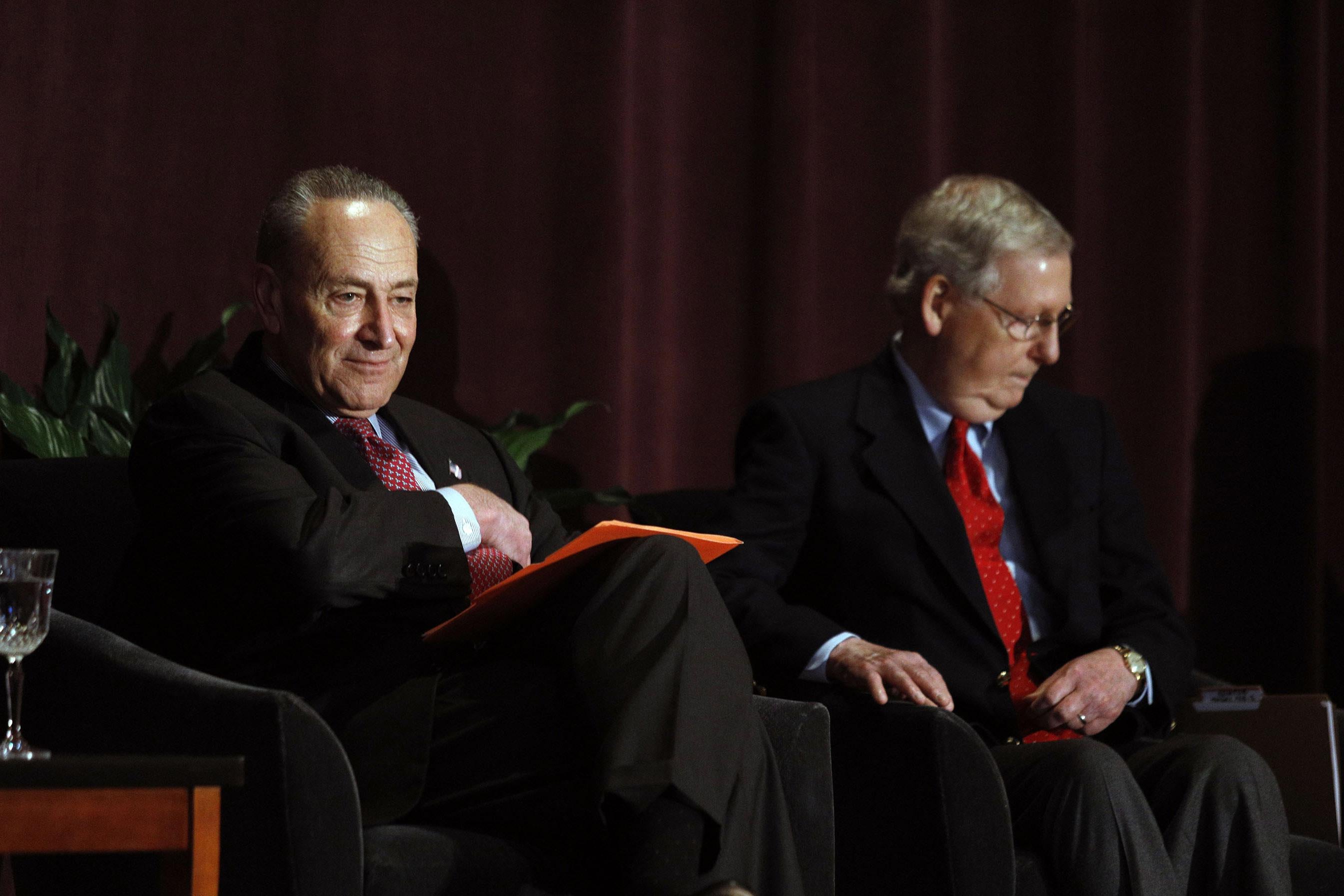 Mitch McConnell and Chuck Schumer sit on a stage in front of plants and a curtain.