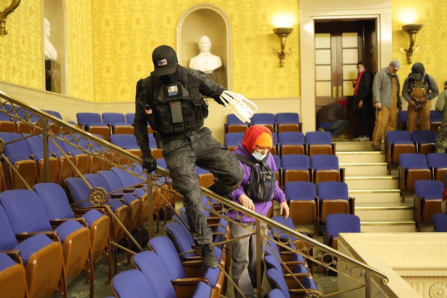 A man wearing black tactical gear and holding zip ties climbs over gallery seats in the Senate chamber.