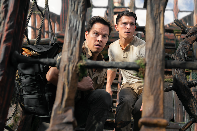 Mark Wahlberg and Tom Holland crouch on a wooden deck, a large wheel nearby.   
